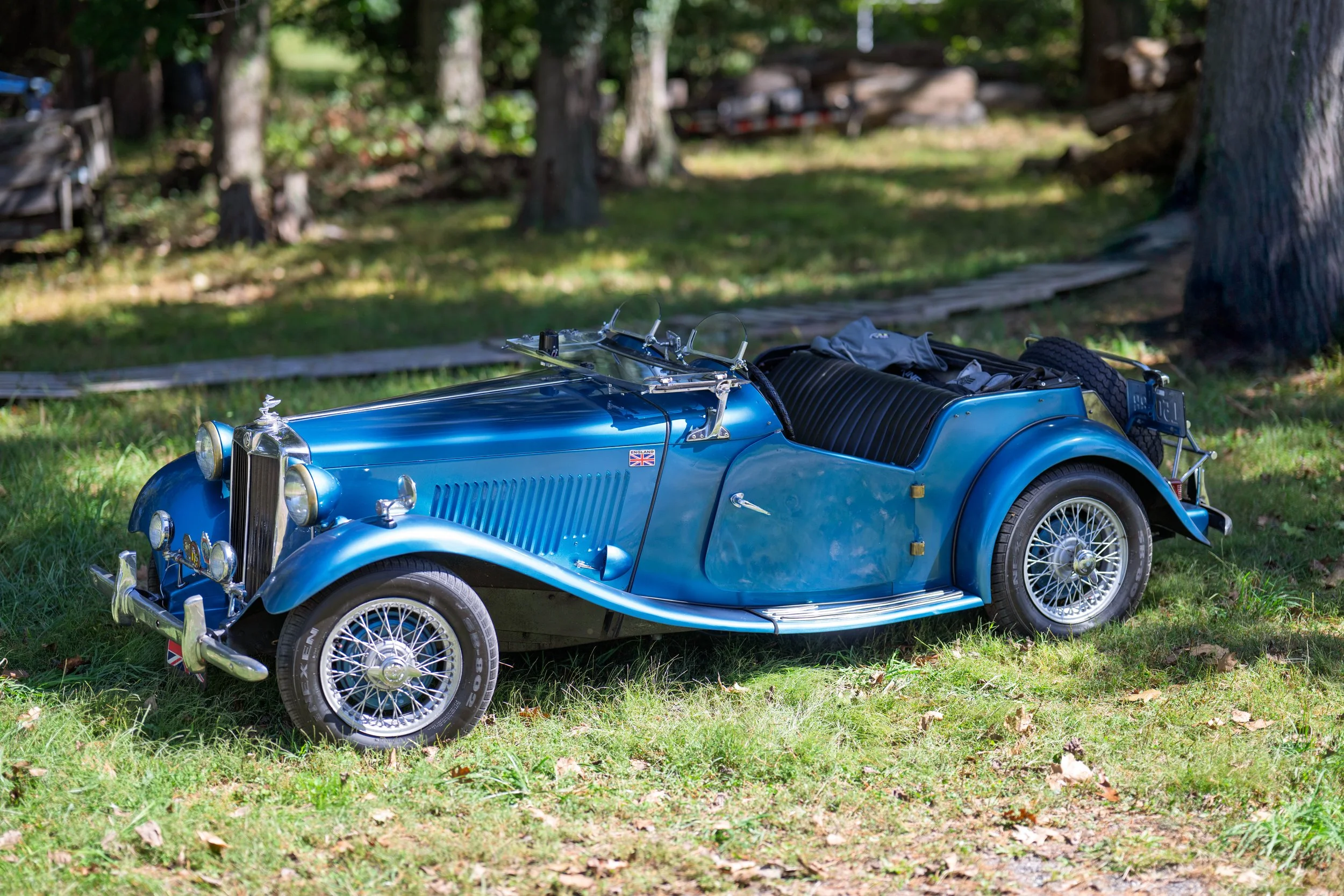 A vintage blue convertible car parked on grass in a wooded area.