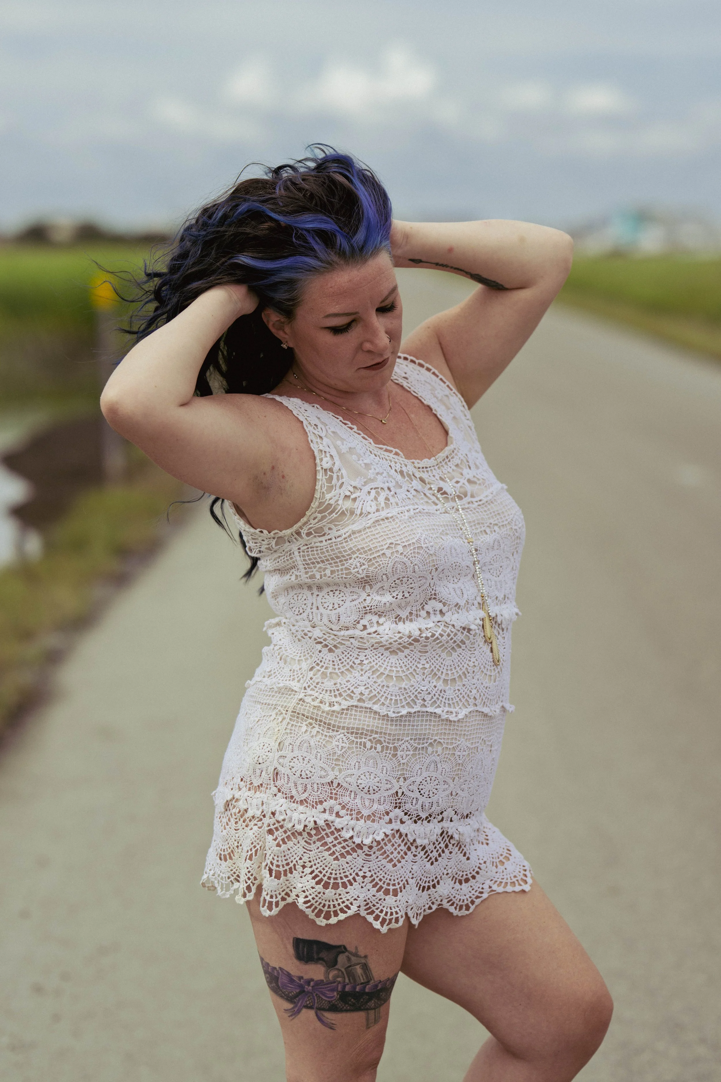 A woman with dark hair streaked with purple, wearing a white lace dress, stands on a rural road with her eyes closed and hands behind her head, outdoors on a cloudy day.