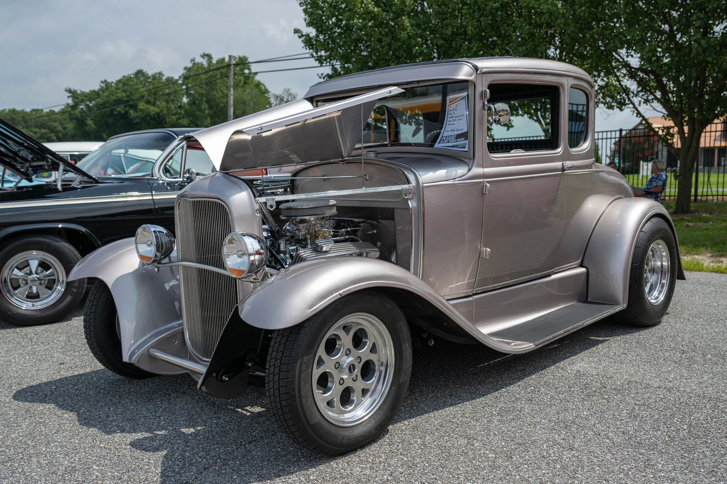 A vintage silver car on display at a car show with its hood open, showing the engine. The car has rounded fenders, chrome details, and a classic design.