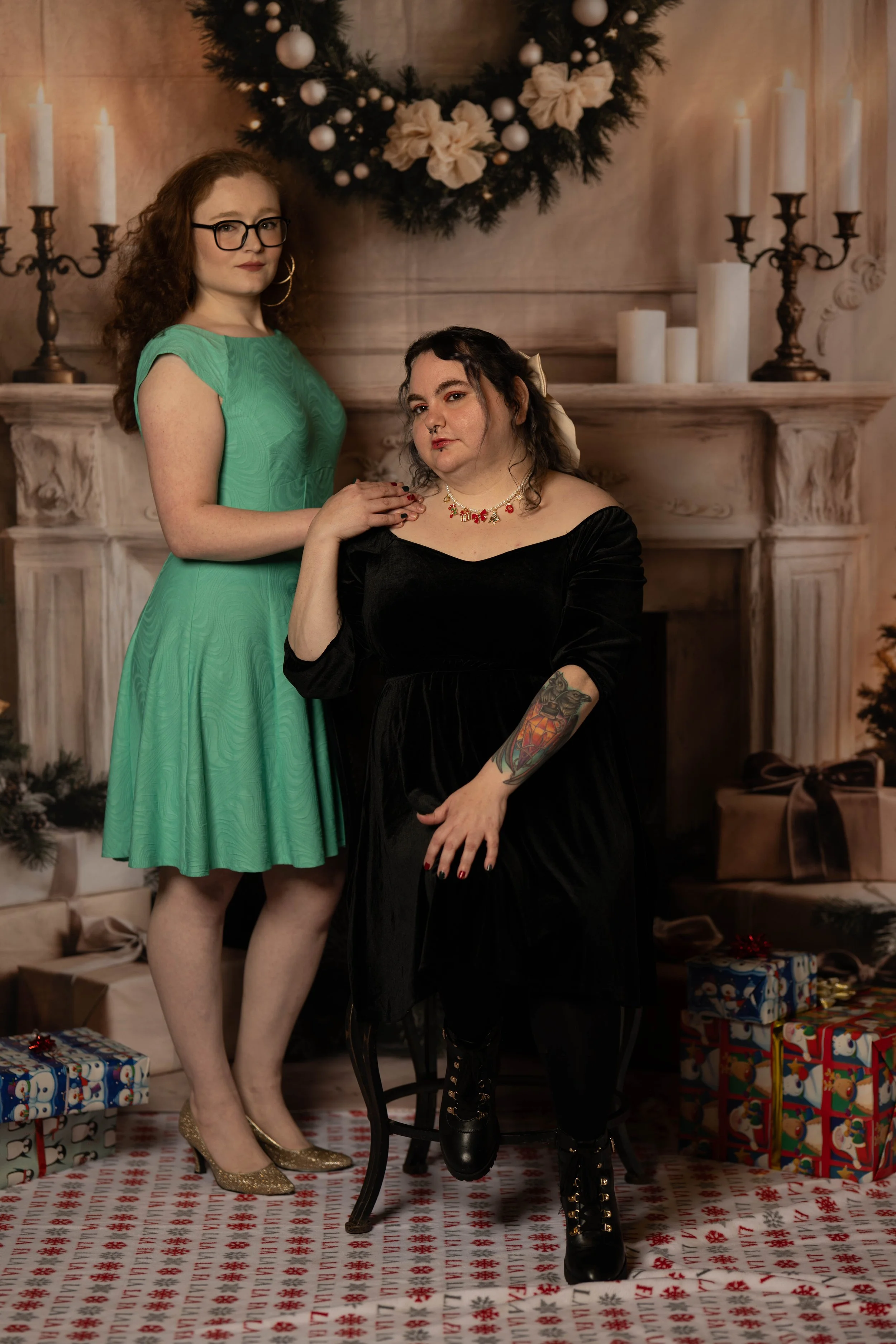 Two women in vintage style posing in front of a Christmas fireplace with stockings, candles, and a wreath. One woman is standing in a green dress with glasses, and the other is sitting in a black dress with tattoos and boots.