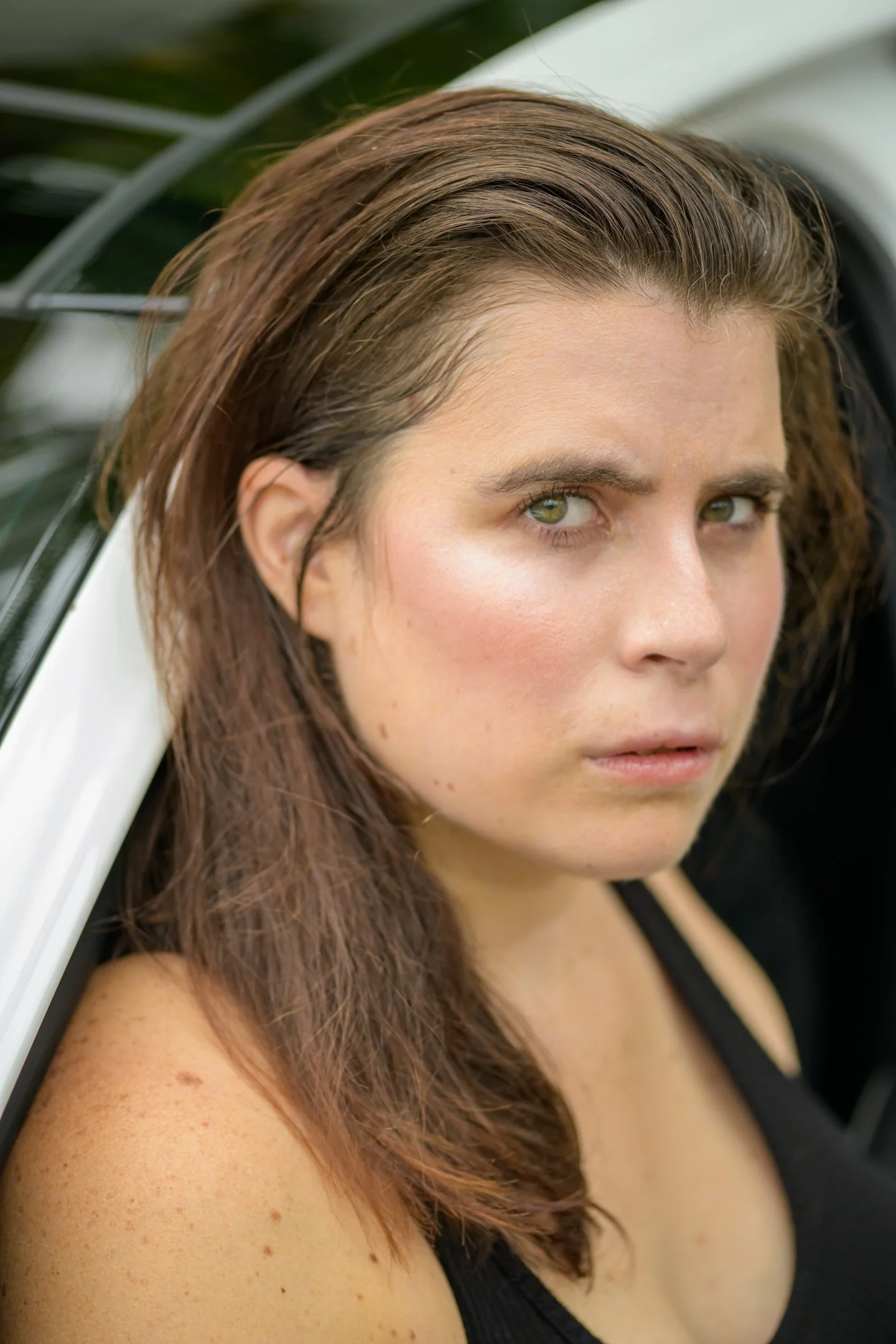 Close-up of a woman with long brown hair and piercing green eyes, looking intensely at the camera, leaning against a white car.
