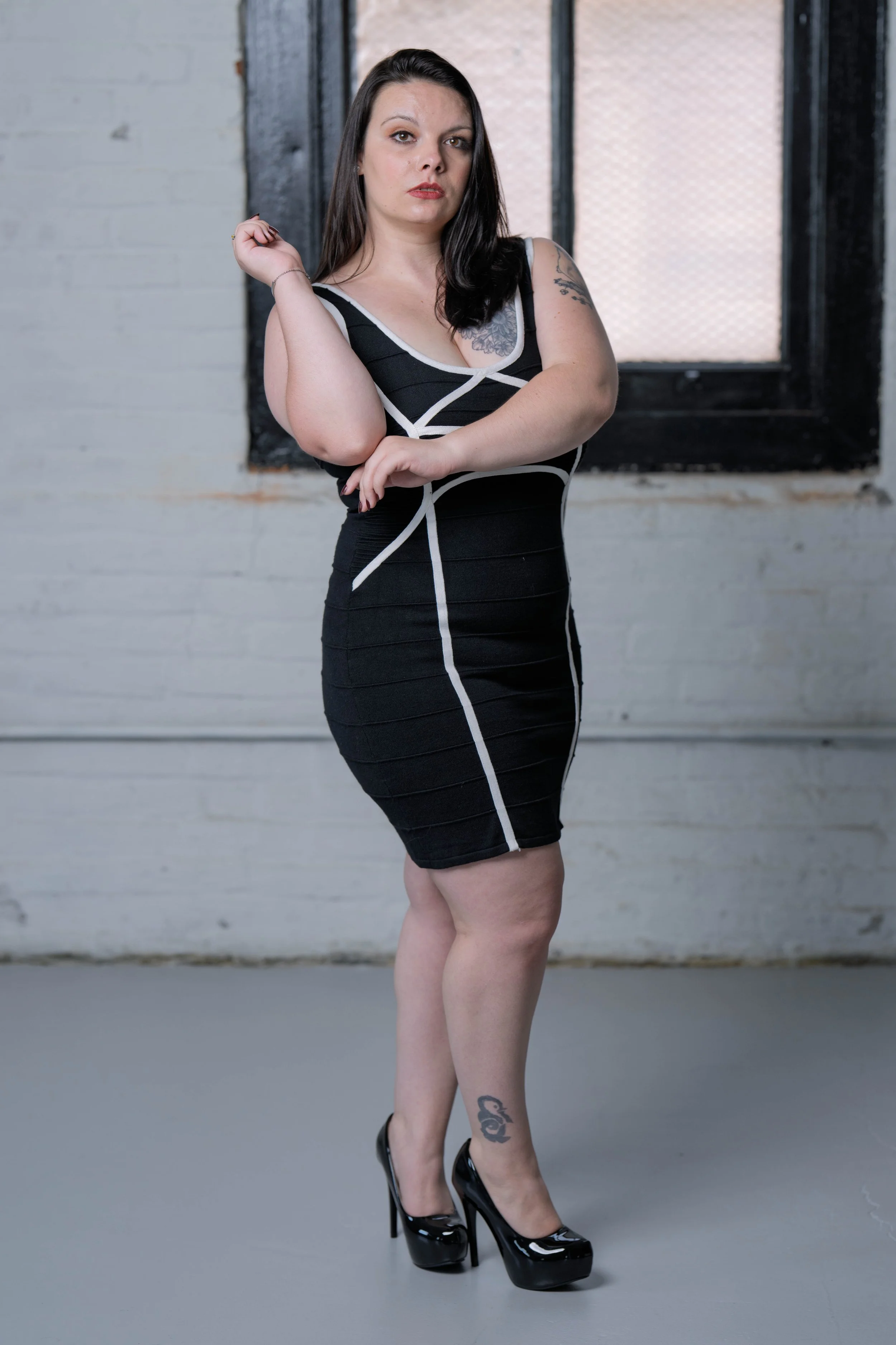 A woman with dark hair wearing a black and white fitted dress and black high heels standing indoors with a white brick wall and a window in the background.