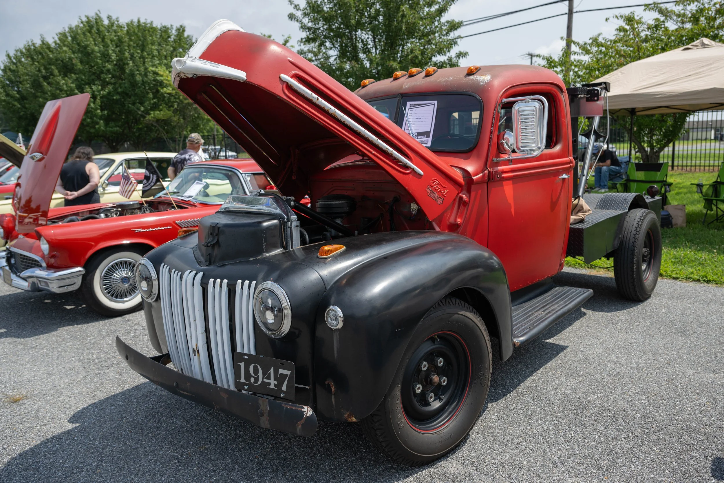A vintage red and black 1947 truck with its hood open at a car show, surrounded by other classic cars and people.