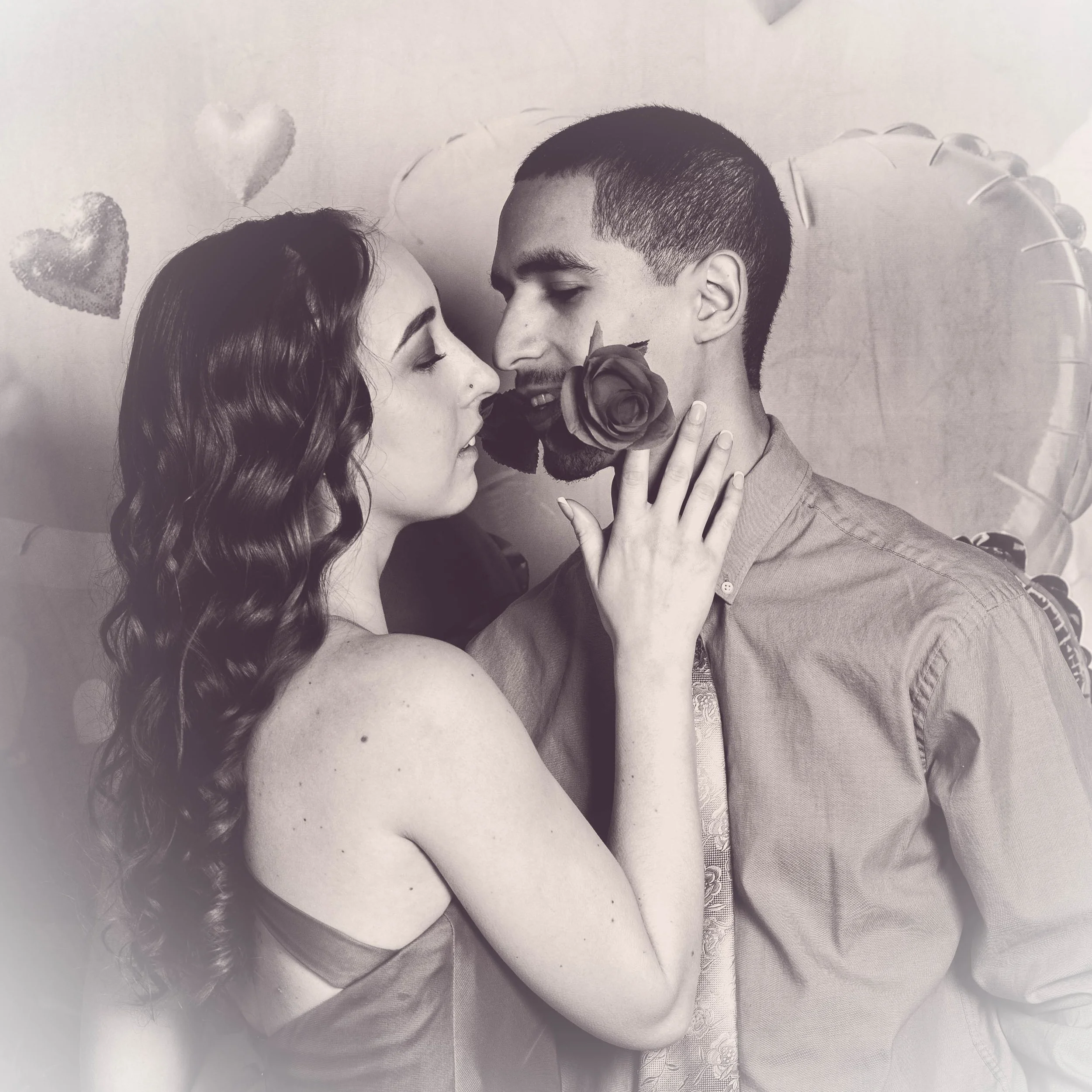 A couple is close together, sharing a romantic moment, with the woman holding a rose near the man's face. They are surrounded by heart-shaped balloons.