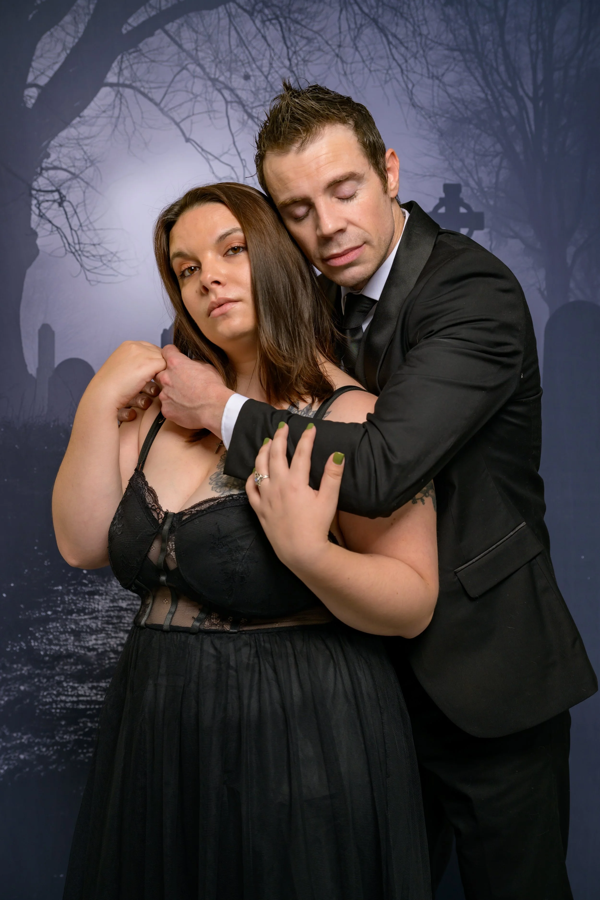 A man in a black suit and a woman in a black dress embrace in front of a spooky, moonlit graveyard backdrop with leafless trees.