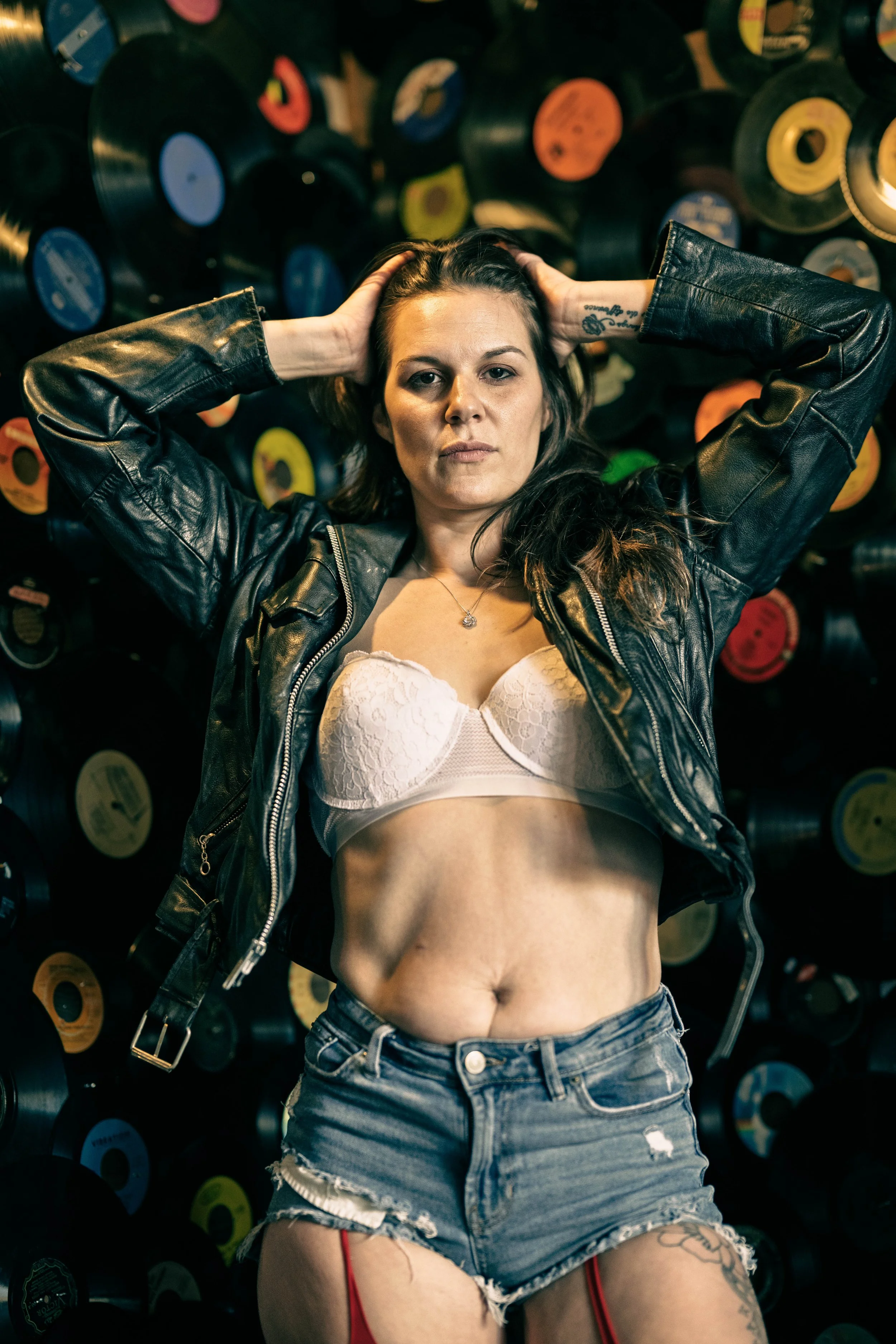 A woman with dark hair in a leather jacket and jeans shorts, wearing a white lace bra, lying on the ground with vinyl records on the wall behind her.