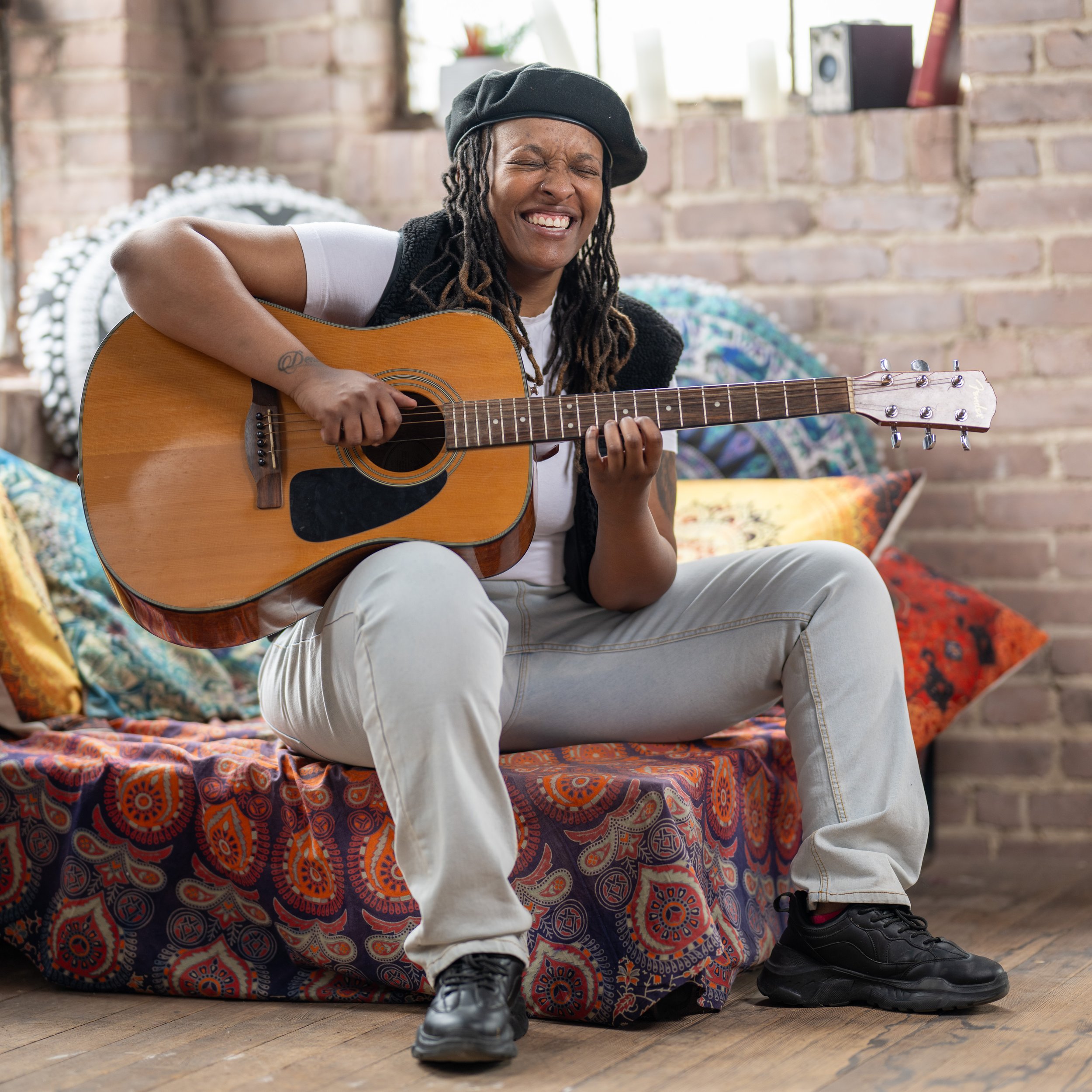 A woman sitting on a colorful bed with pillows, playing an acoustic guitar, smiling with eyes closed, in a cozy room with a brick wall background.