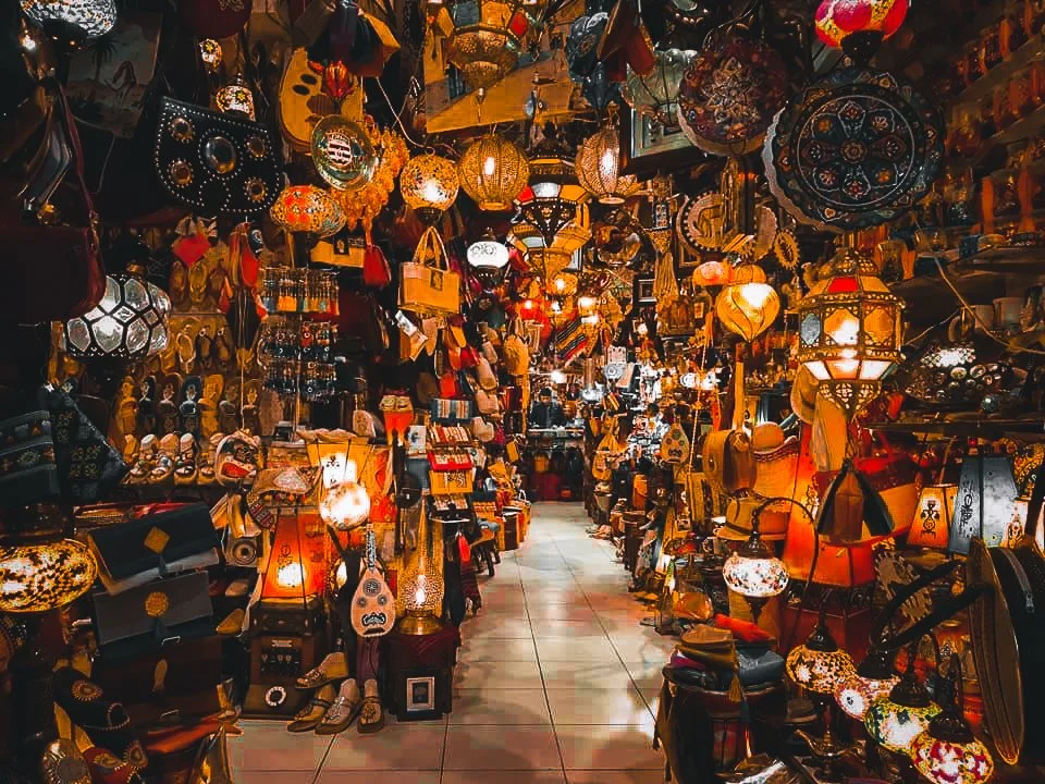 A narrow marketplace aisle filled with colorful hanging and table lamps, decorative items, and accessories in warm, vibrant lighting.