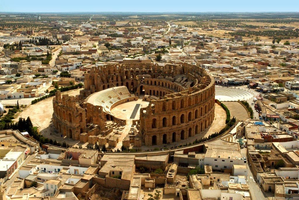Aerial view of the ancient Roman Colosseum in a city, with surrounding buildings and streets visible.