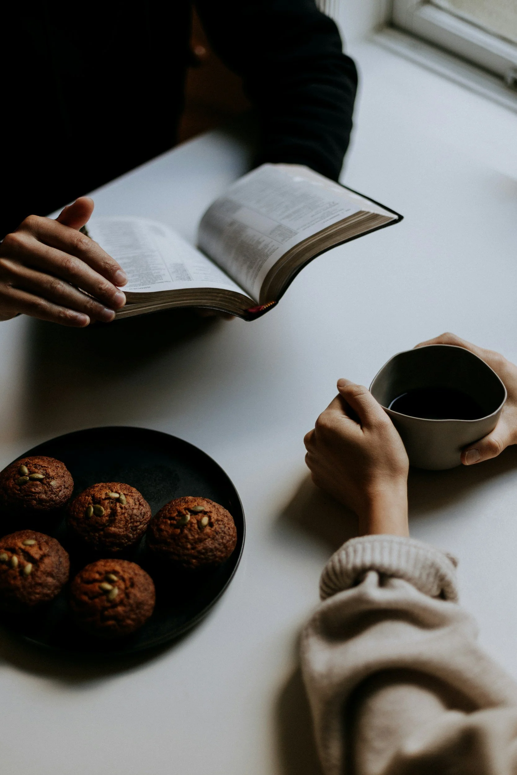 Two people sitting at a white table, one holding a mug of coffee and the other reading a Bible. There is a plate of six muffins on the table near the person with the mug.