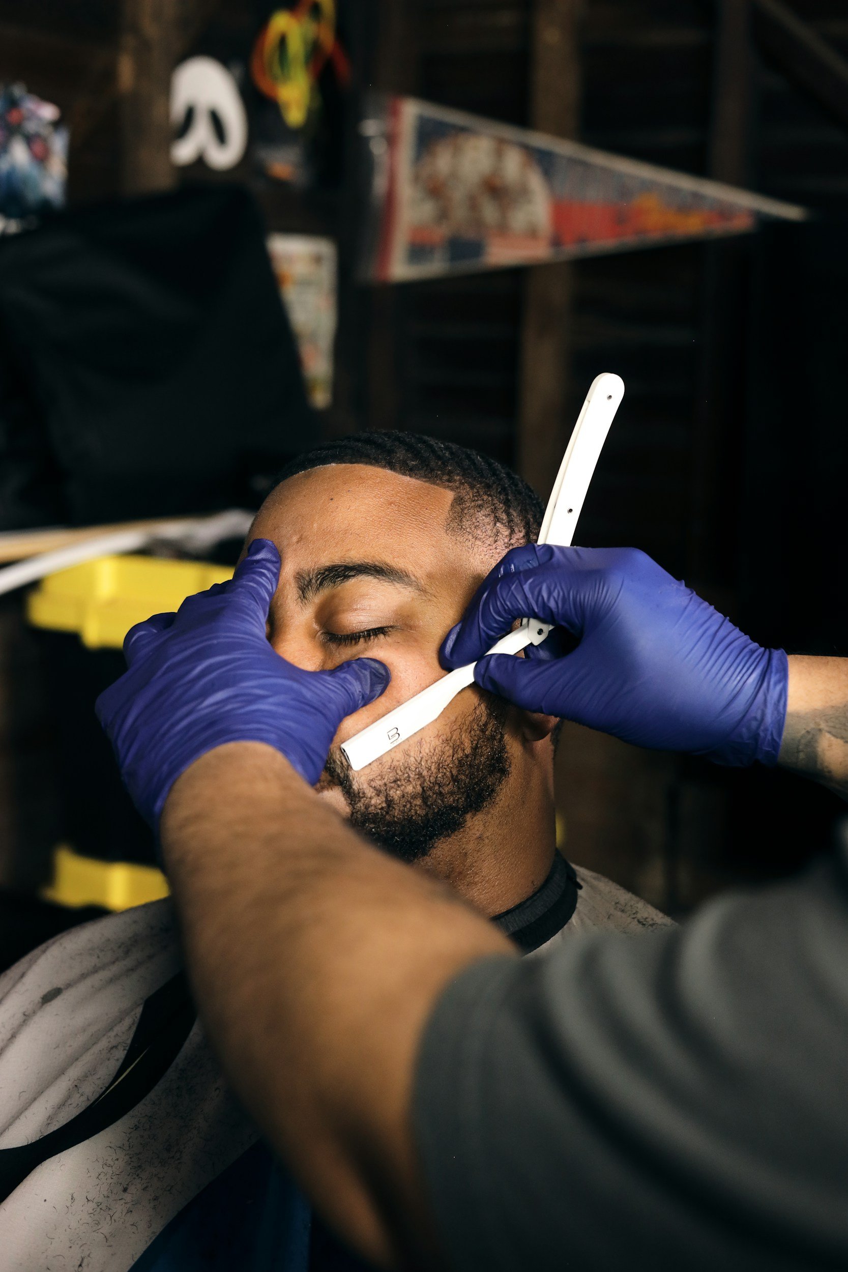 A man is getting a close-up tattoo applied to his face by a tattoo artist wearing blue gloves, in a tattoo studio with dark wood walls and tattoo-related artwork in the background.