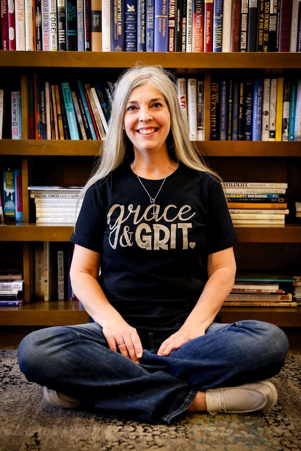 A woman with long gray hair sitting cross-legged on an area rug in front of a bookshelf, smiling at the camera. She is wearing a black t-shirt that says 'grace & grit' and a necklace with a cross pendant.