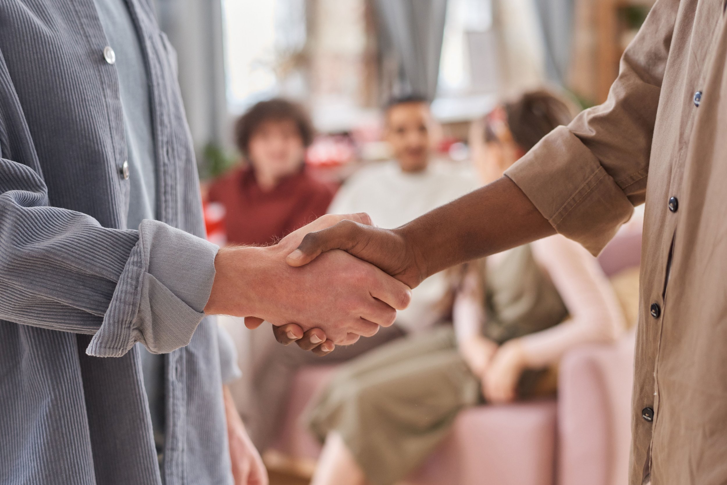Two people shaking hands at a meeting or event, with a group of smiling individuals in the background.