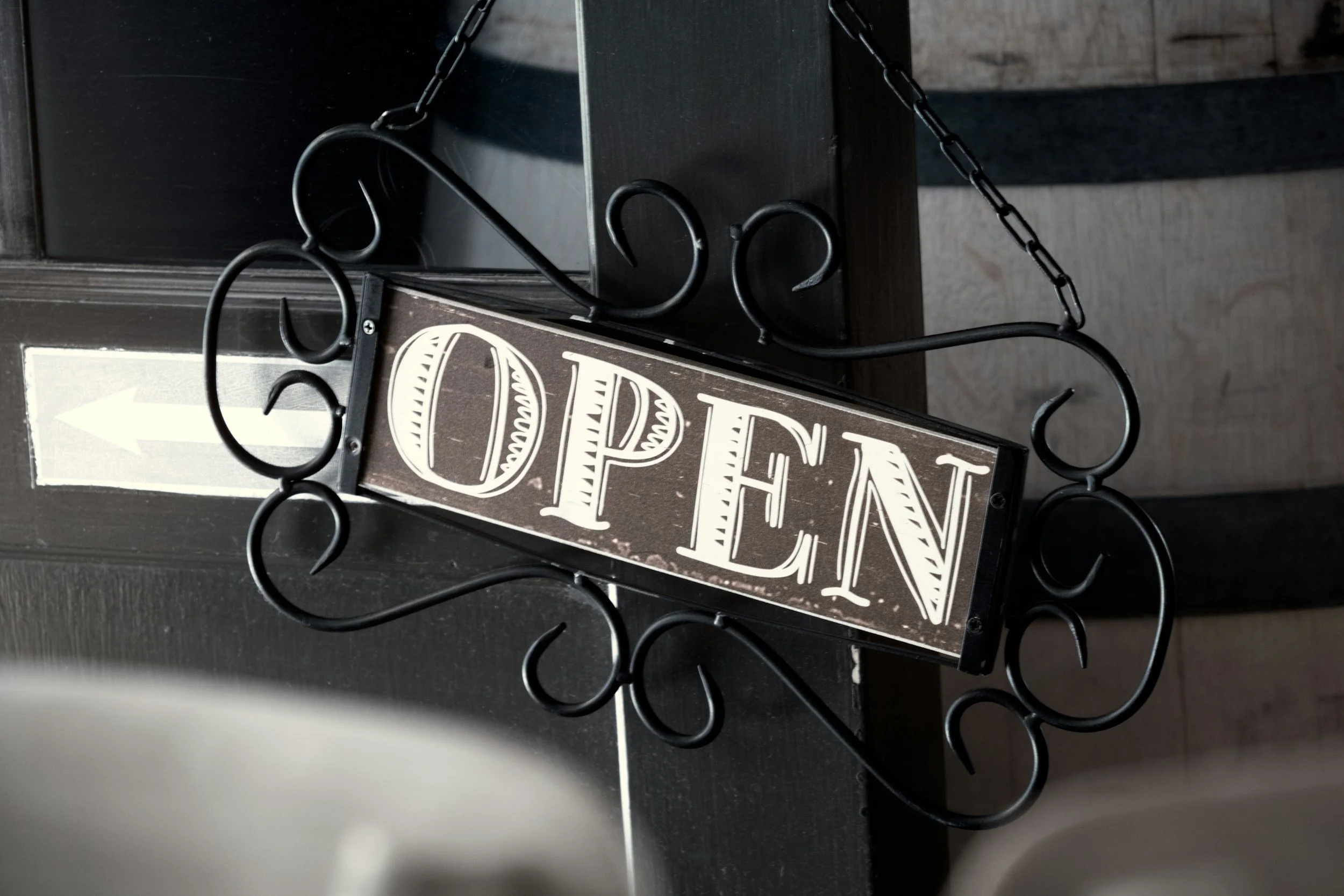A decorative black metal sign featuring the word 'OPEN' in white letters, hanging from a chain on a black surface.
