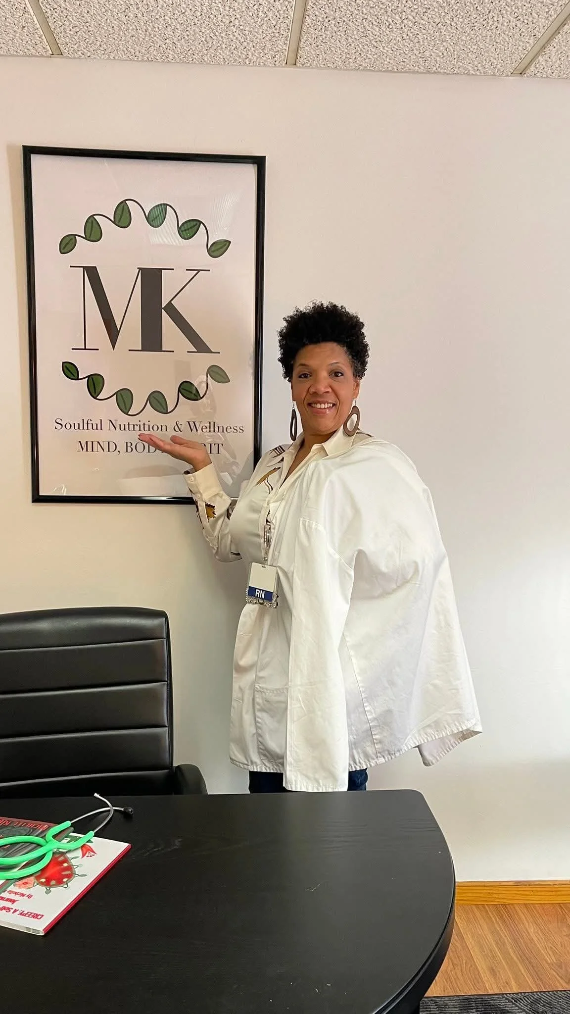A woman in a white coat standing next to a sign that reads "MK Soulful Nutrition & Wellness MIND, BODY, SPIRIT" and pointing to it.