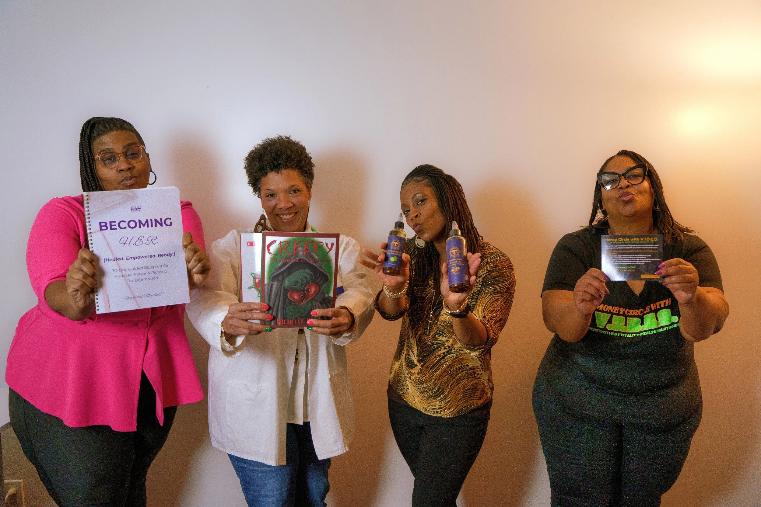 Four women standing against a plain wall, holding various books and products, smiling and posing for the camera.