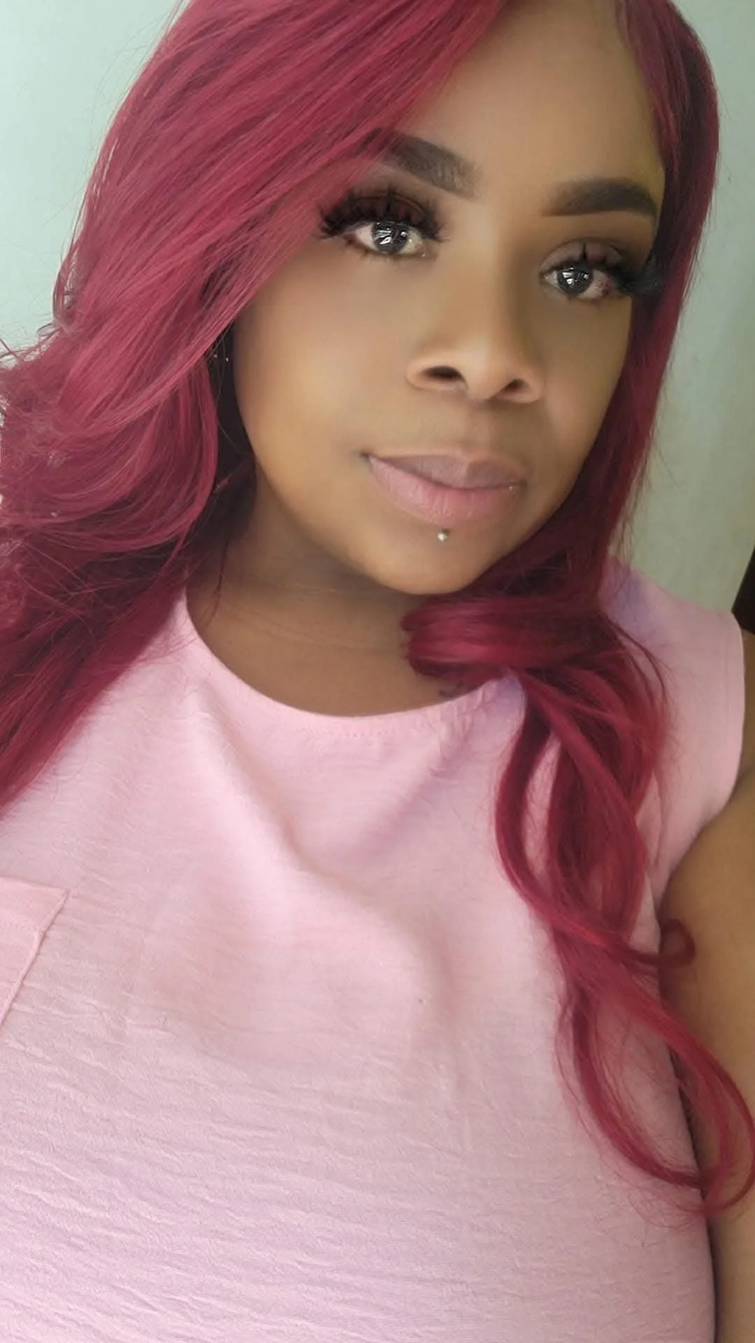 A woman with long, wavy pink hair, wearing a light pink top, with makeup and a small piercing below her lips, taking a selfie.