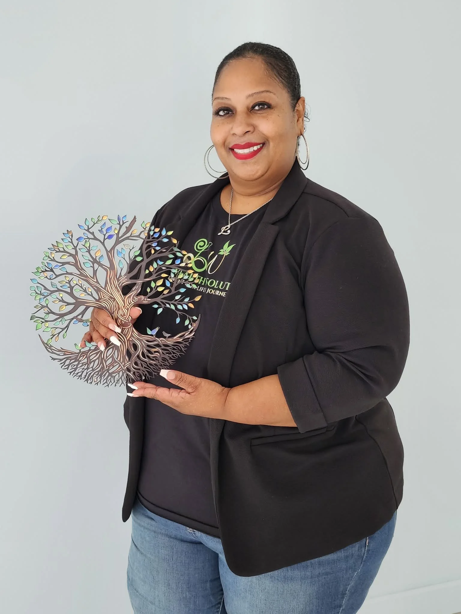A smiling woman with glasses poses with her arms raised, standing in front of a mirror in an indoor space. She is wearing a black and green patterned button-up shirt and dark pants.