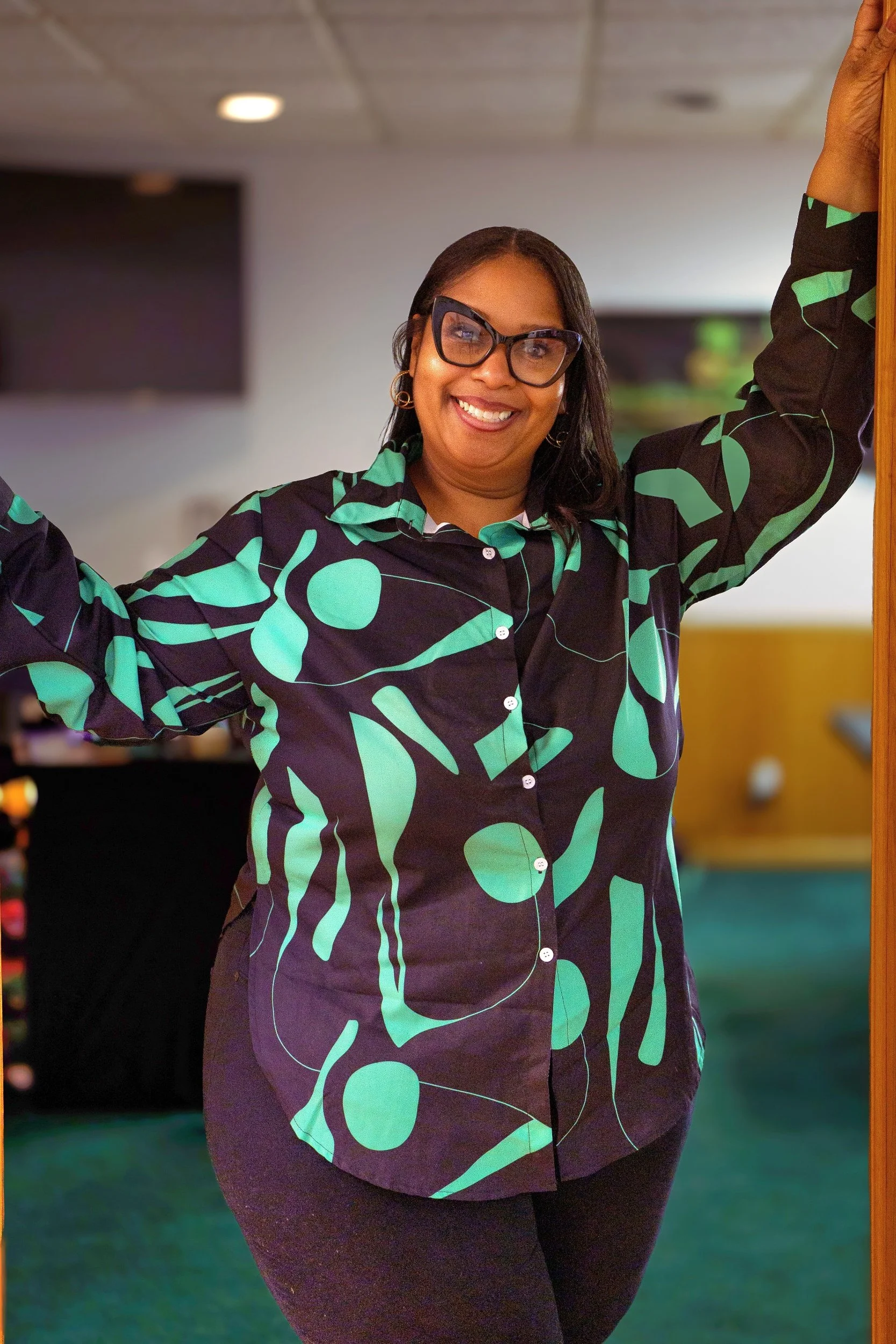 A smiling woman with glasses poses with her arms raised, standing in front of a mirror in an indoor space. She is wearing a black and green patterned button-up shirt and dark pants.