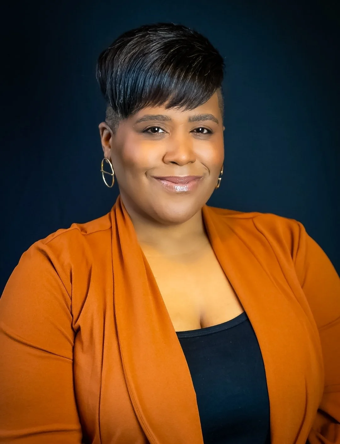 A woman with short black hair, wearing hoop earrings, a black top, and an orange blazer, smiling against a dark background.