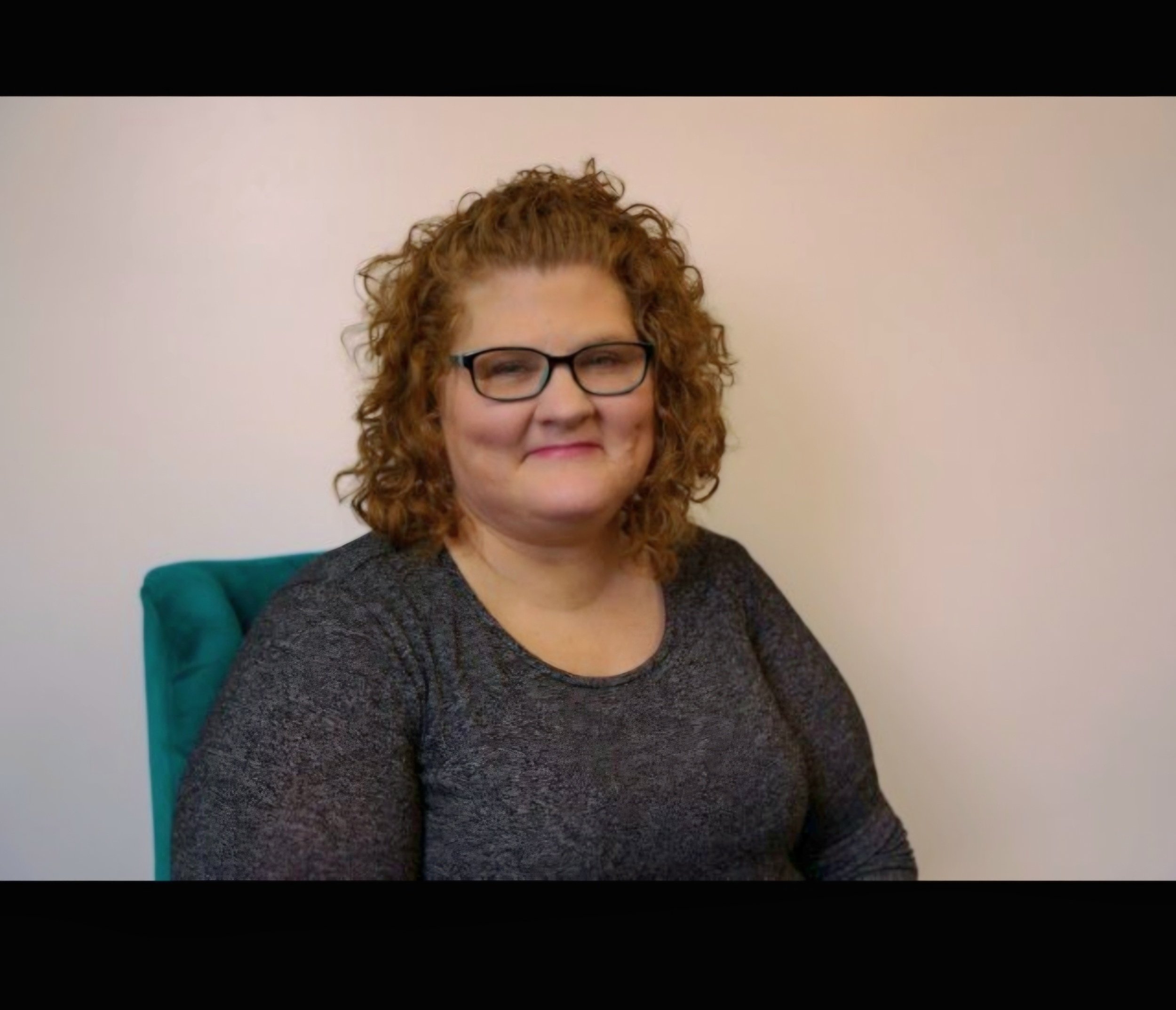 A woman with curly red hair, glasses, and a gray long-sleeve shirt, sitting in a teal chair against a plain light-colored wall.