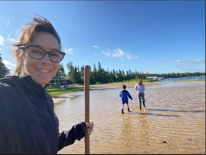 Chantal Banda with glasses smiling while holding a stick on a sandy beach, with her two children walking in the water near trees and boats in the background on a sunny day.