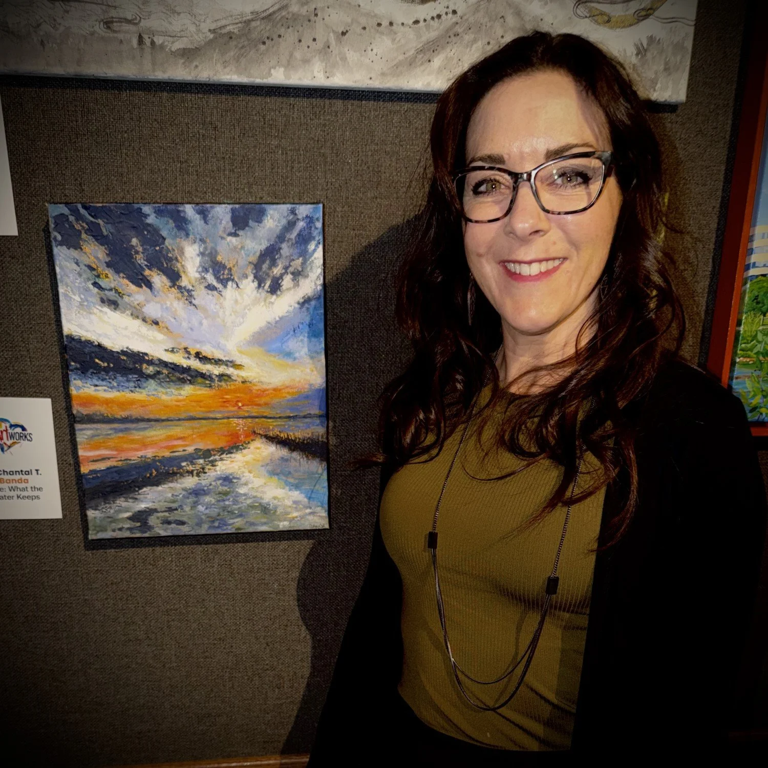 A woman with glasses and long brown hair smiling at an art gallery, standing next to a colorful landscape painting of a sunset over water with dramatic clouds.