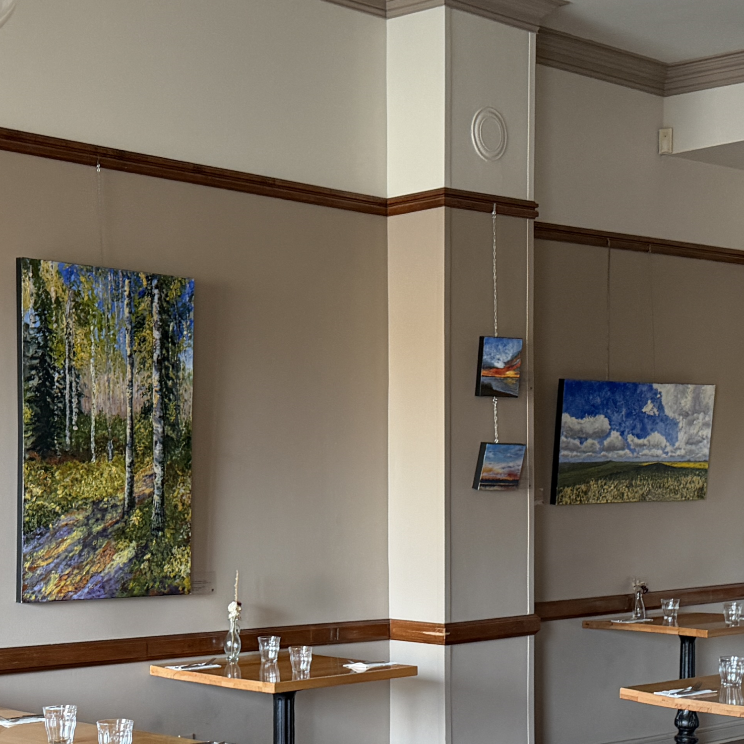 Interior of an art gallery showcasing landscape paintings, including a forest scene, a sunset, and clouds over a field. Tables are set with glasses and silverware.