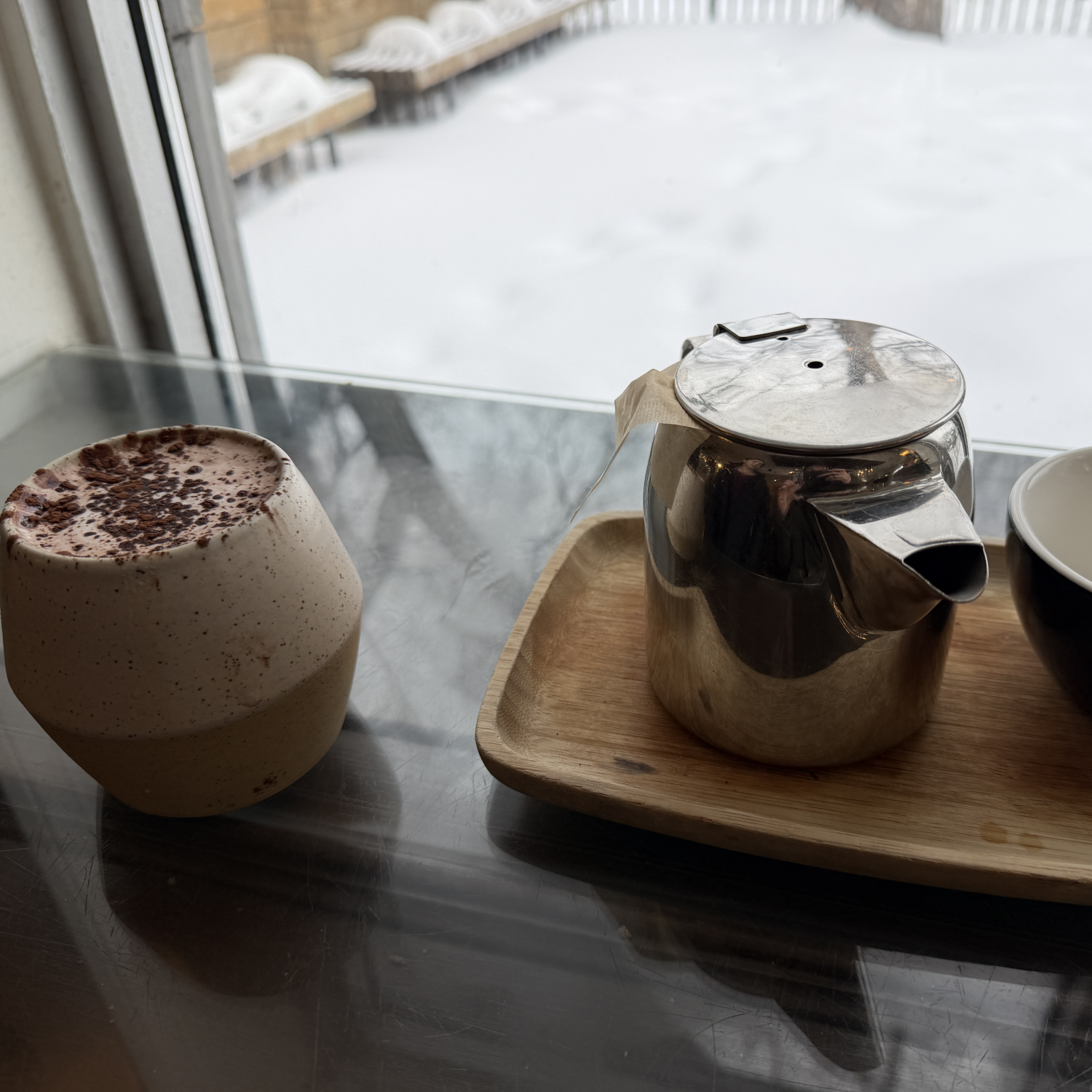 A ceramic mug filled with a frothy hot chocolate topped with chocolate shavings, a metallic teapot on a wooden tray, and part of a black and white bowl on a dark reflective table near a window with snow outside.