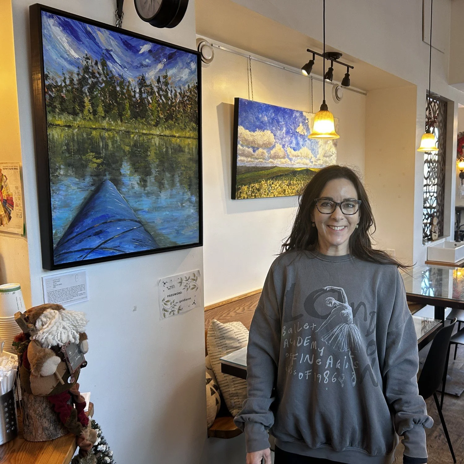 Chantal Banda, an artist, is standing in an art gallery. Two of her landscape paintings hang on the wall behind her, one depicting a lake scene with trees and the other a sky with clouds over fields.