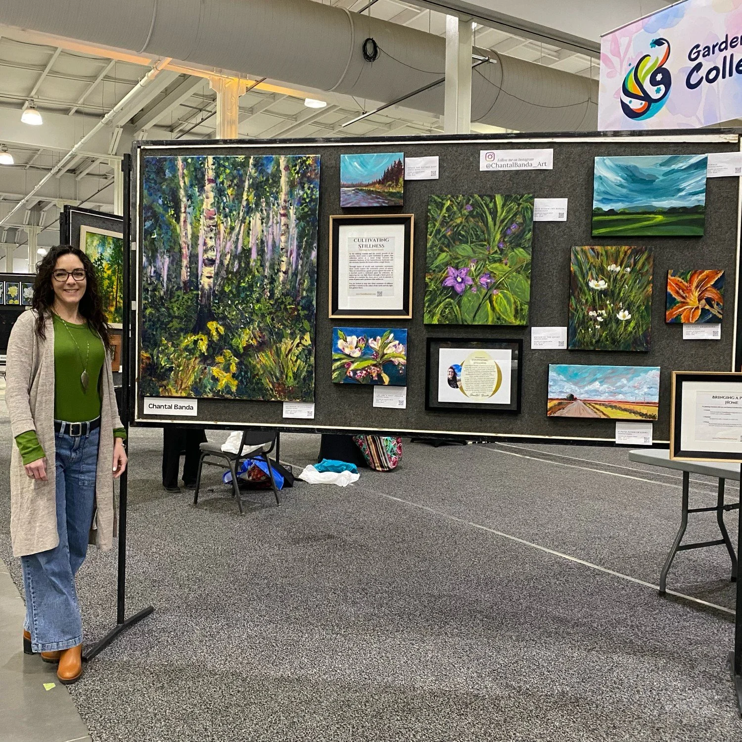 Chantal Banda with dark curly hair, glasses, a green shirt, jeans, and a long beige cardigan standing next to an art display at an indoor art show. The display features various landscape and floral paintings arranged on a black fabric-covered panel.