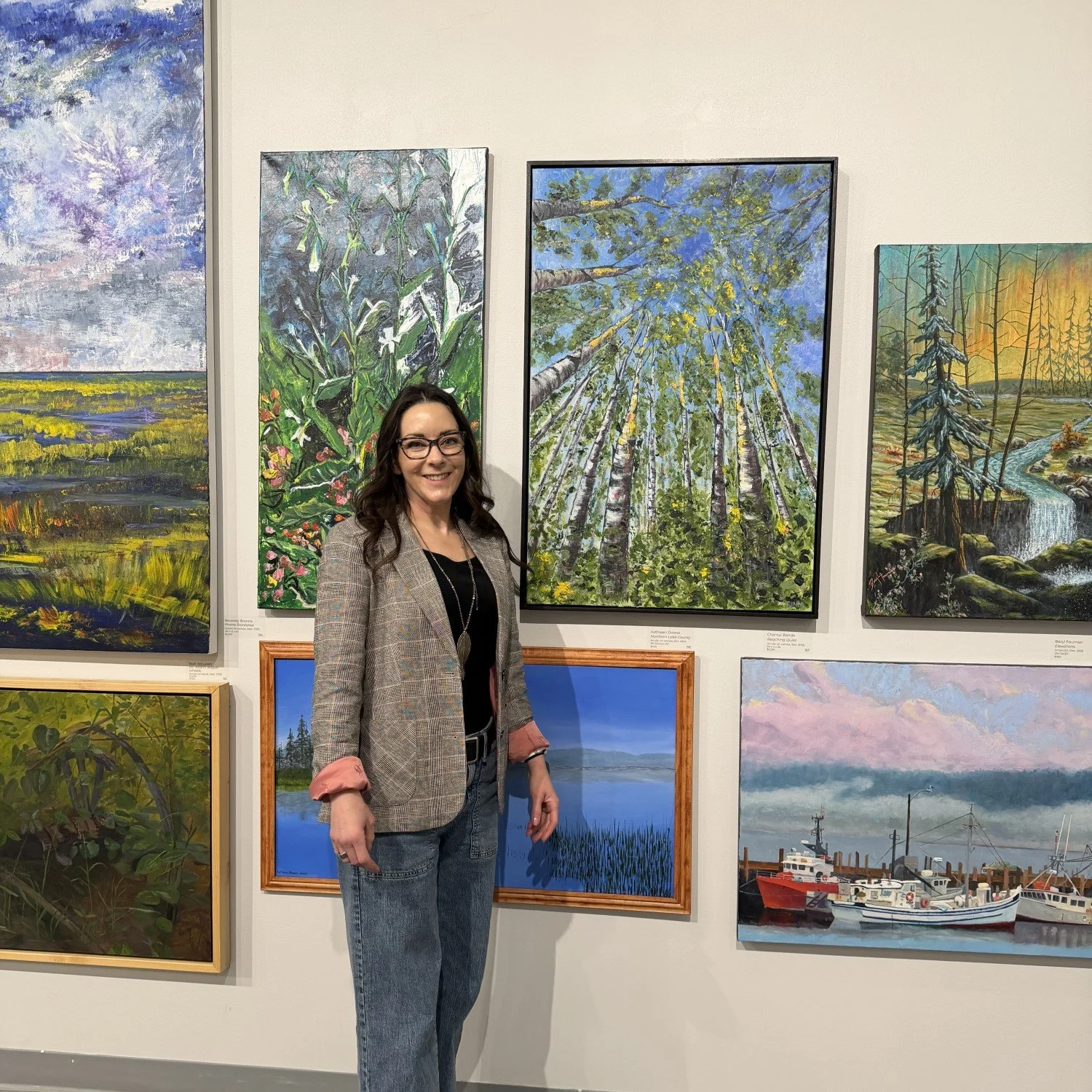 Chantal Banda with long dark hair, glasses, wearing a plaid blazer and jeans, standing in an art gallery in front of several landscape paintings, smiling at the camera.