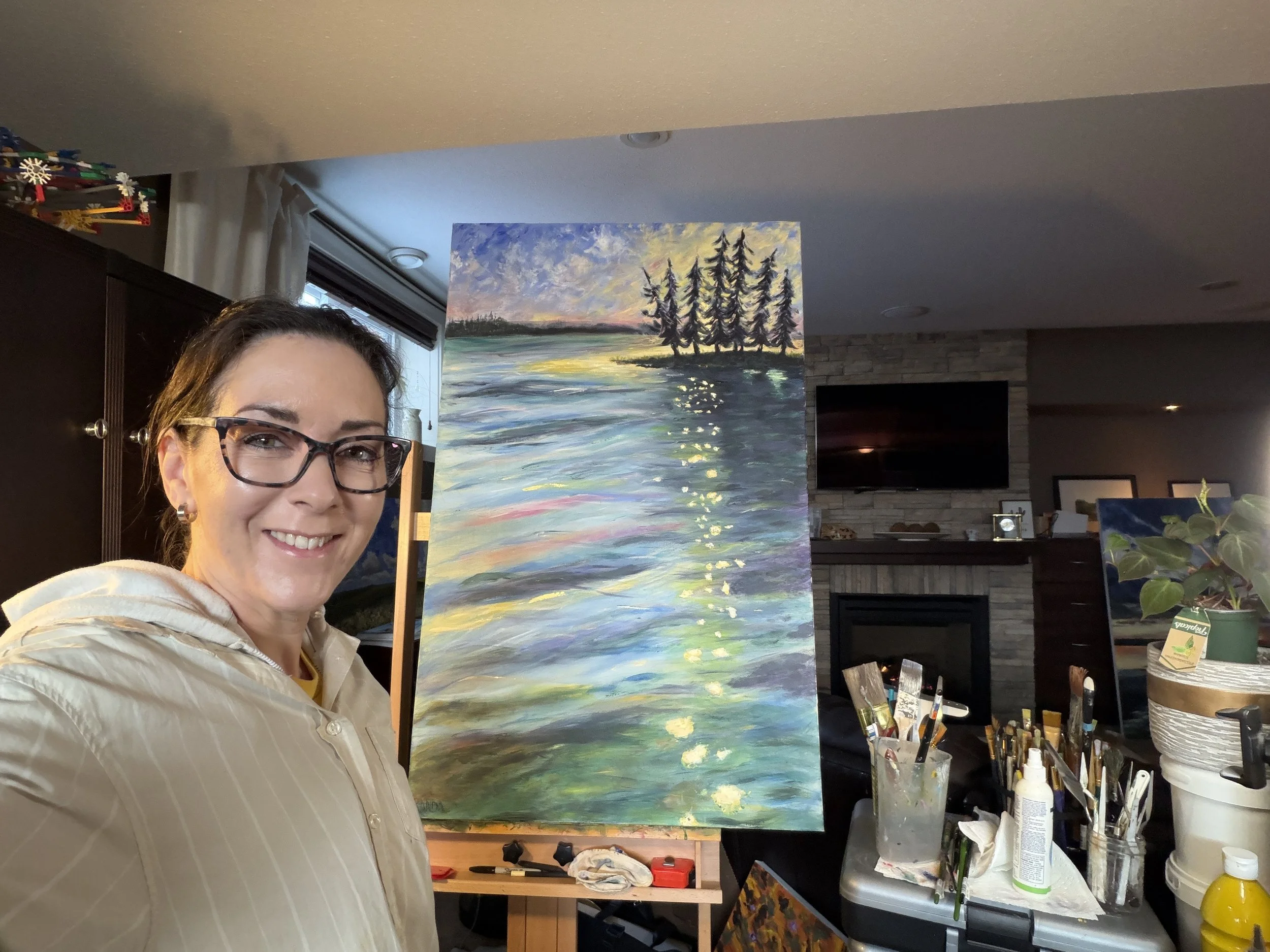 Chantal Banda smiling with glasses in an art studio, standing next to an impressionist-style painting of water, trees, and a sunset.