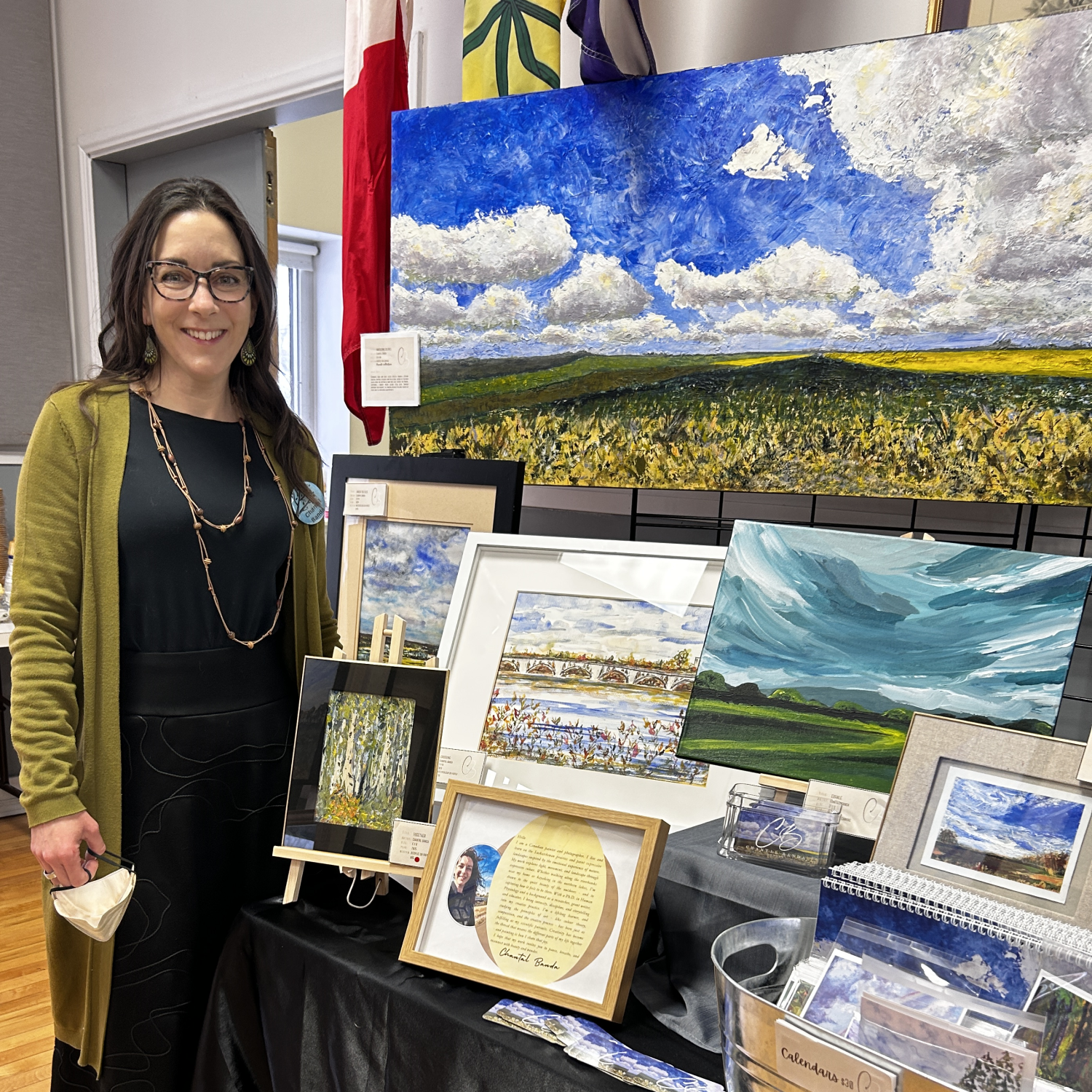 Chantal Banda standing next to a display of landscape paintings at an art exhibit. The paintings feature scenes of skies, fields, and waterways. The woman is smiling, wearing glasses, a black dress, and a yellow cardigan.