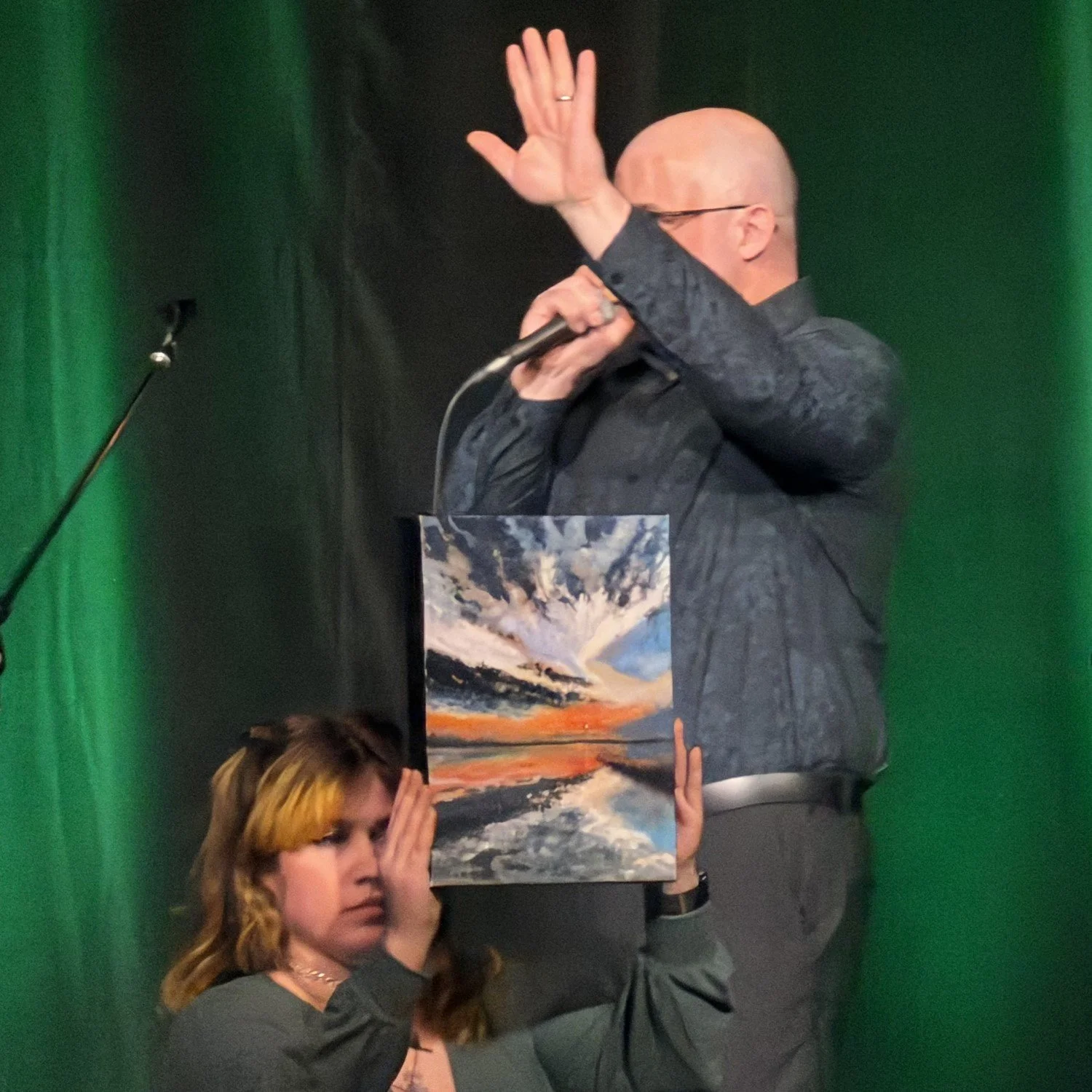 A man with glasses, dressed in a dark shirt, is speaking into a microphone with his hand raised at an art auction. A woman with red hair, wearing gray, holds a colorful landscape painting up in front of her face.