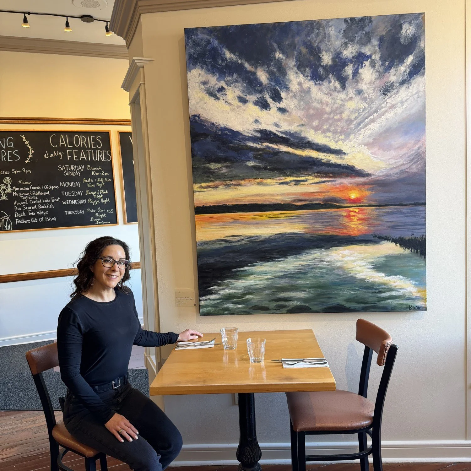 Chantal Banda, an artist, is sitting at a restaurant table. Behind her is her large landscape painting of a sunset over water with clouds in the sky.