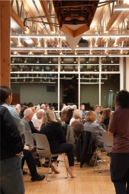 Large group of people attending a literary book conference or presentation in a modern, well-lit room with floor-to-ceiling windows.