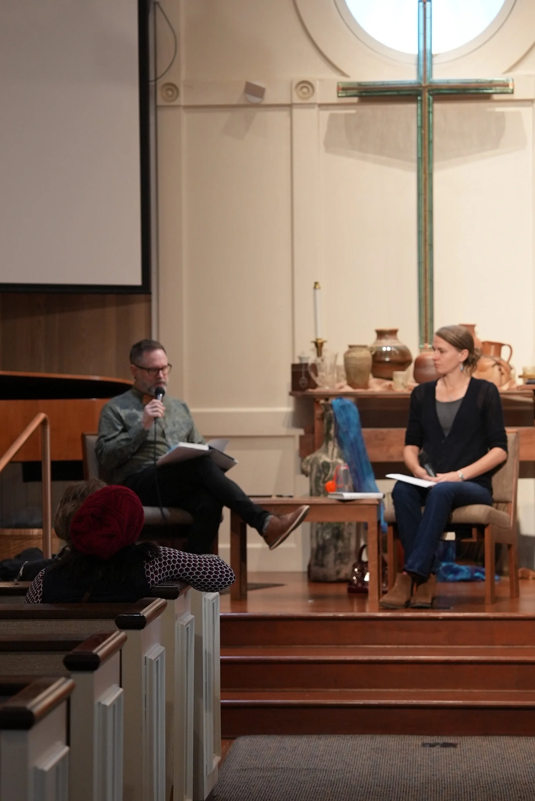 Two people seated on a stage in a church or religious setting, with a large cross behind them, engaged in a discussion or presentation. One person is holding a microphone and reading from a book, while the other has a notepad and a microphone in hand