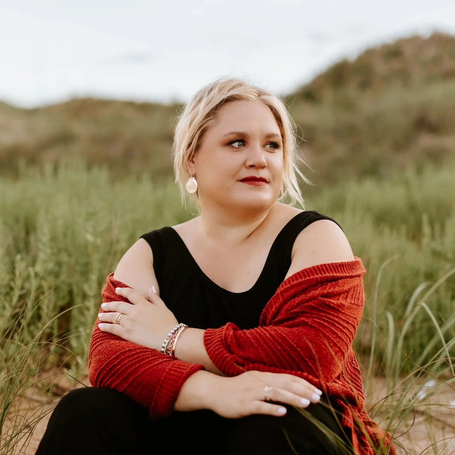 A woman with blonde hair and earrings sitting outdoors among tall grass, wearing a black top and a red sweater draped over her shoulders, looking to the side.
