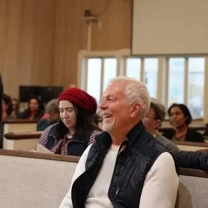 A smiling elderly man with white hair and a beard sitting in a church or similar setting surrounded by diverse people, including a woman wearing a red beret.
