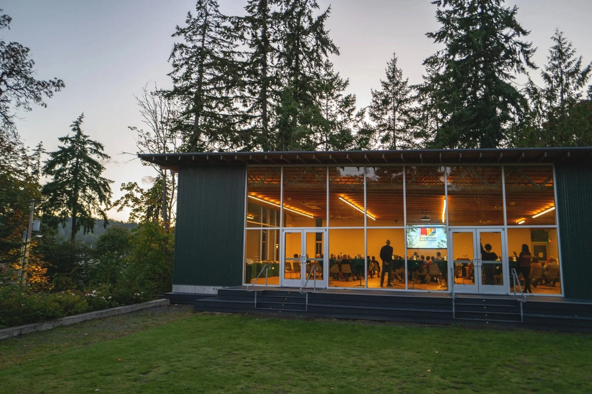 A modern building with large glass windows revealing an indoor literary event with people seated and an author standing at the front. The building is set amidst tall trees in a lush outdoor area during dusk on Bainbridge Island, Washington.