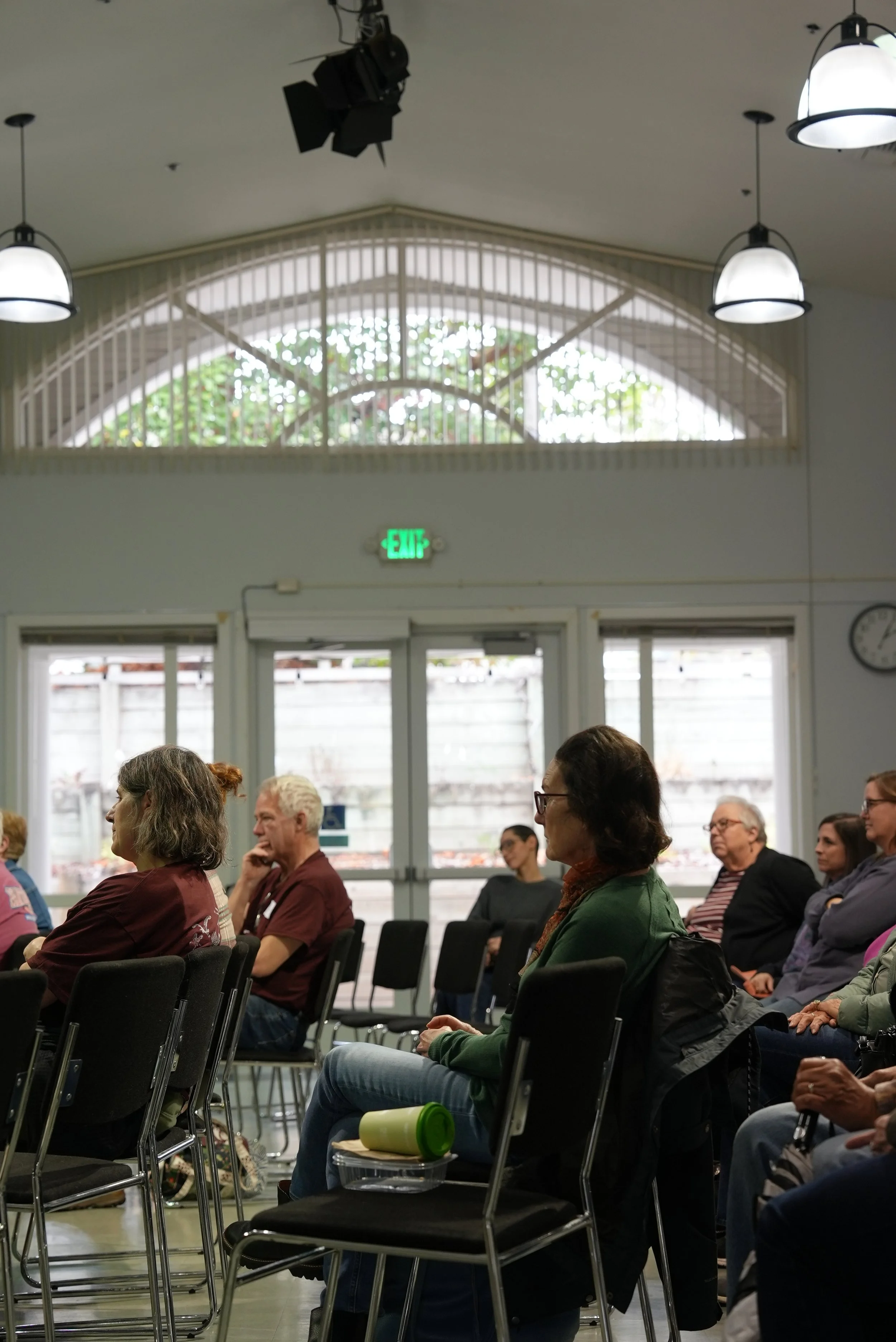 A group of people sitting and listening at an event in an indoor space with large windows and ceiling lights.