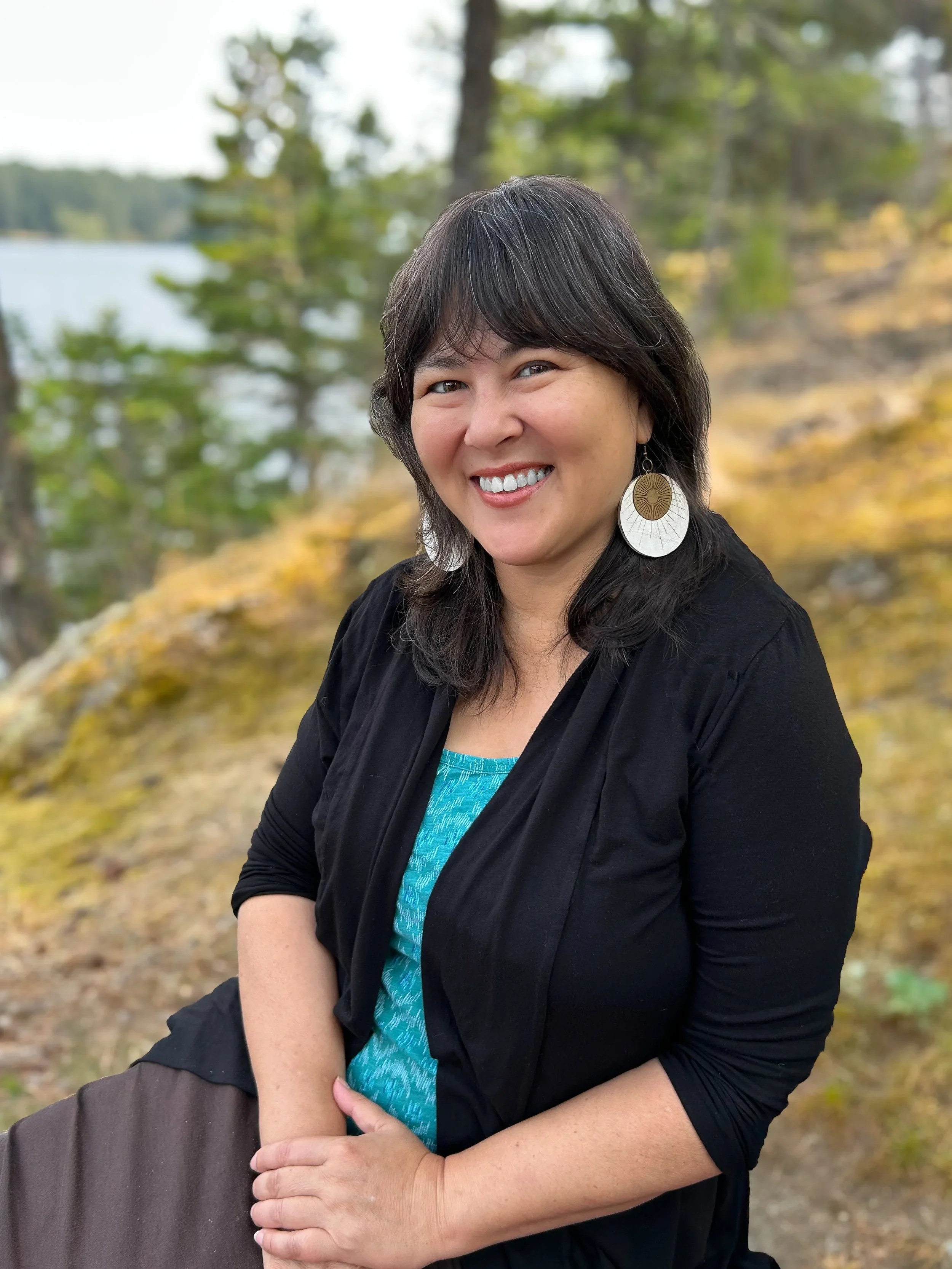 A smiling woman with shoulder-length dark hair and large circular earrings, wearing a black jacket and a blue top, standing outdoors with trees and water in the background.