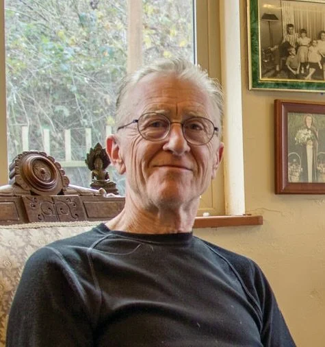 An elderly man with glasses and gray hair sitting in a cozy room with framed photos on the wall behind him.