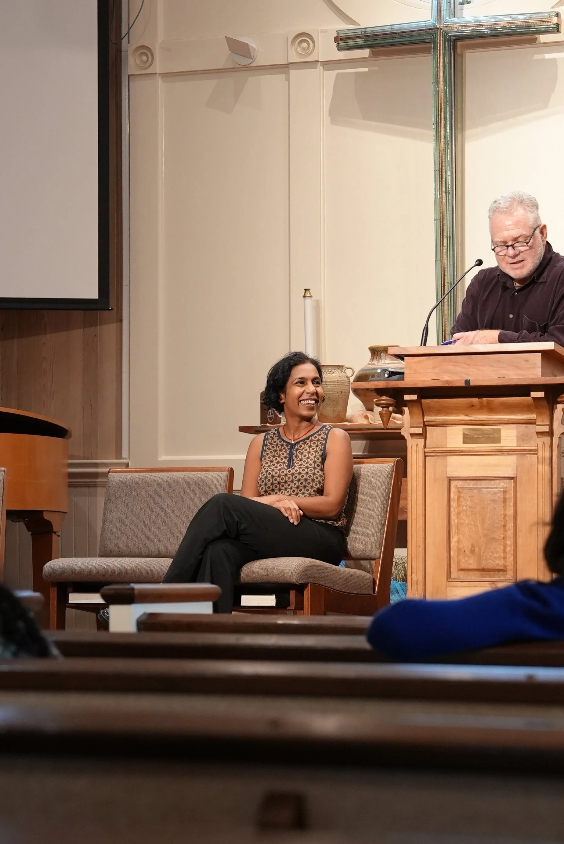 A woman author sitting on a chair, smiling, at a public speaking event or lecture, with a man standing at a wooden podium with a microphone in front of her, in a room with a large cross on the wall.
