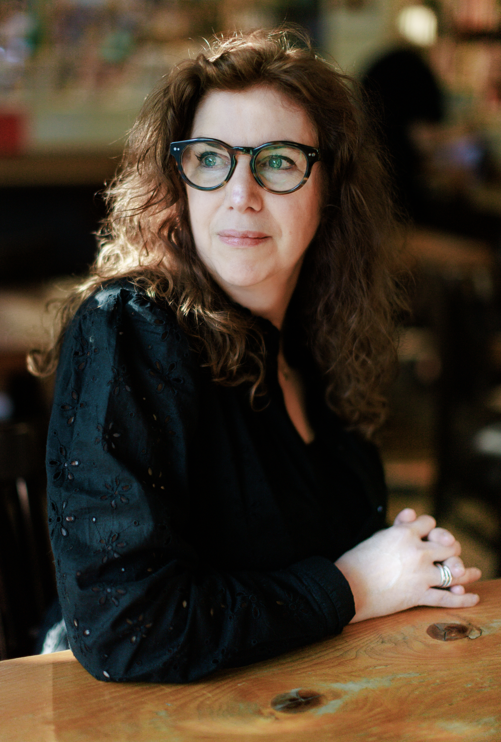 A woman with curly brown hair and glasses sitting at a wooden table in a cozy, dimly-lit indoor space.