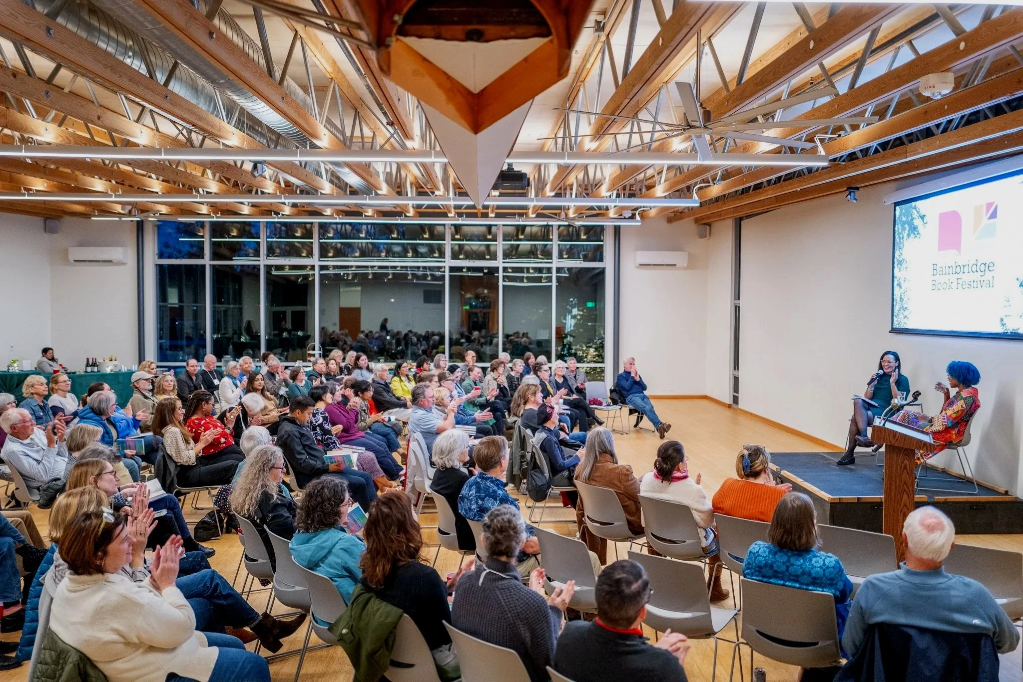 An indoor event with a large audience seated and listening to two women authors speaking on stage. The room has wooden beams and large windows at the back, with a projector screen displaying the Bainbridge Island Book Festival logo.