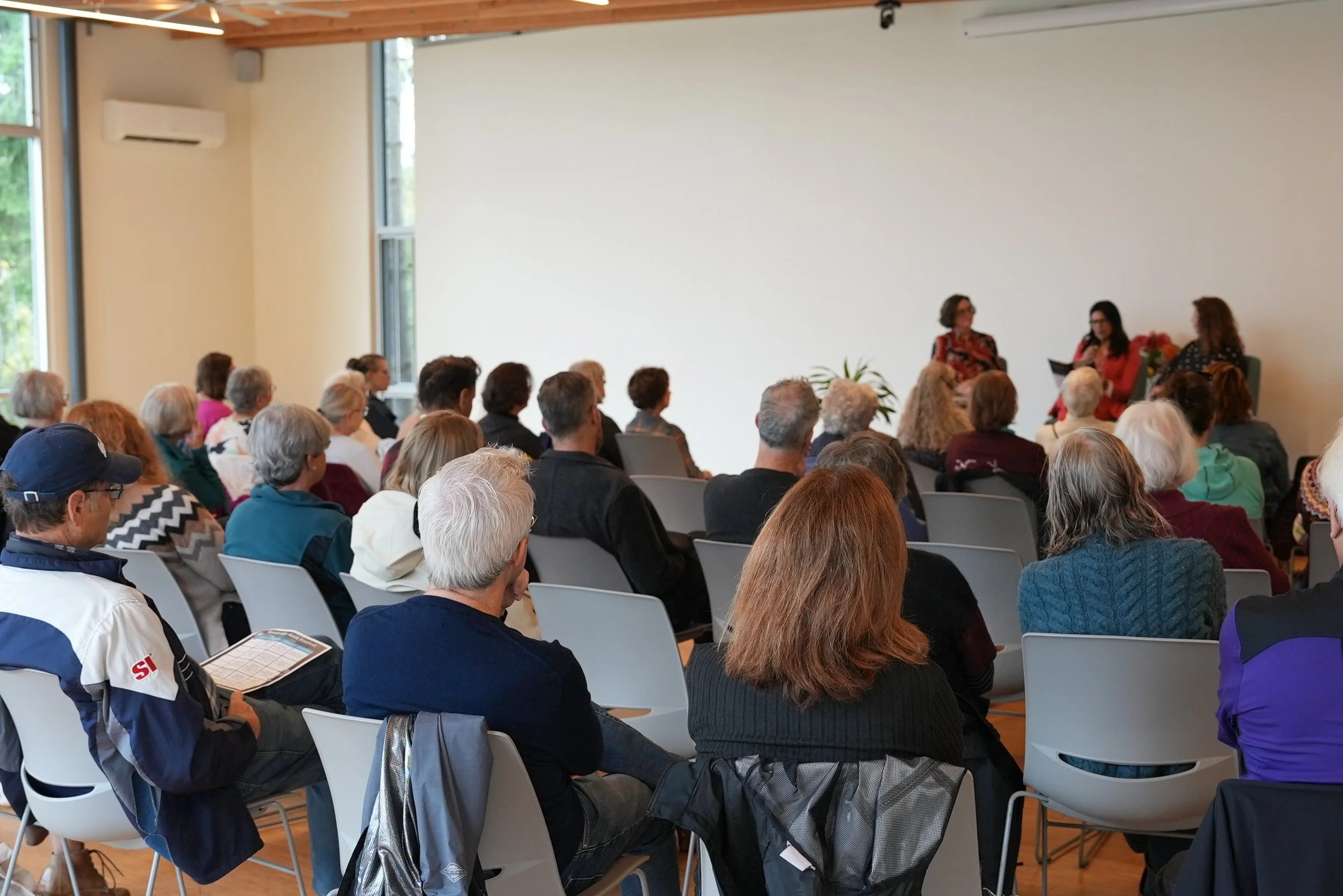 A large group of people seated and listening to three women writers speaking at the front of a room during a presentation or panel discussion.