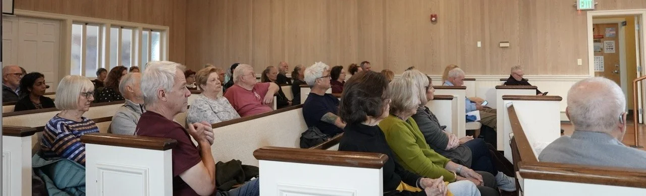 People seated inside a church or community hall, facing forward, listening to a speaker or presentation about books.
