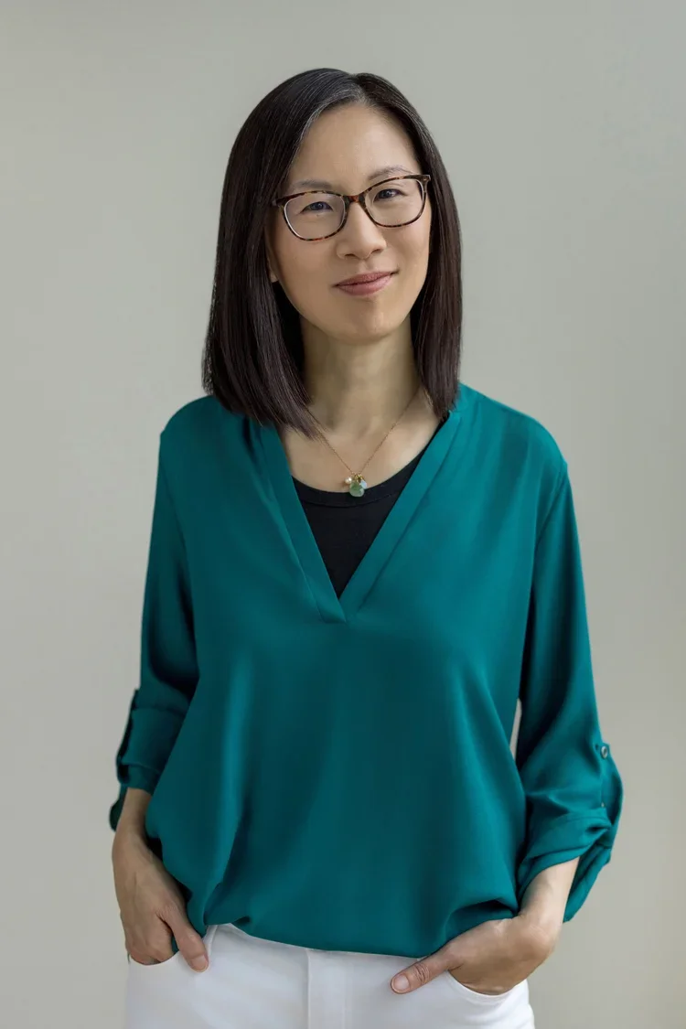 A woman with shoulder-length black hair, wearing glasses, a green blouse, and white pants, standing against a plain light gray background.